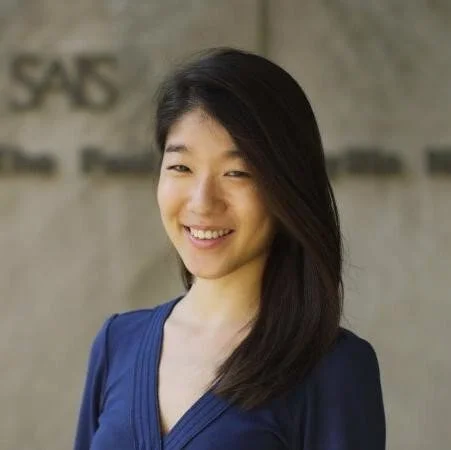 Young woman with long dark hair smiling, wearing a blue top, standing outdoors in front of a beige wall with some text.