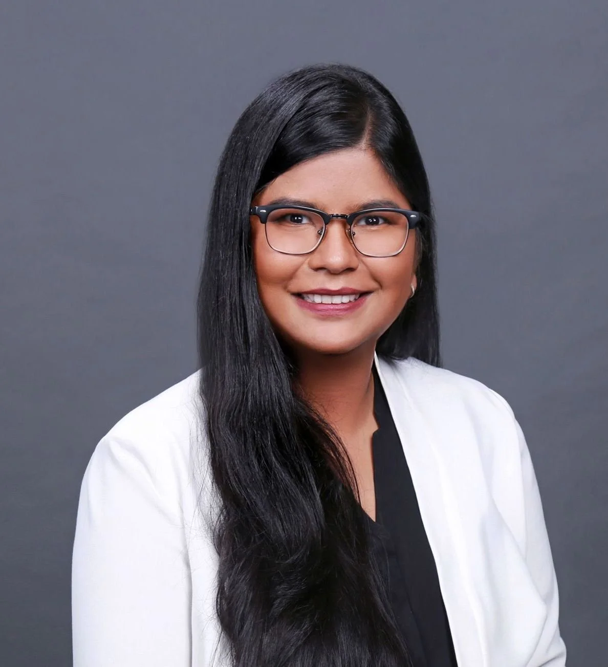 Professional headshot of a woman with long black hair, glasses, smiling, wearing a white blazer and black top against a gray background.