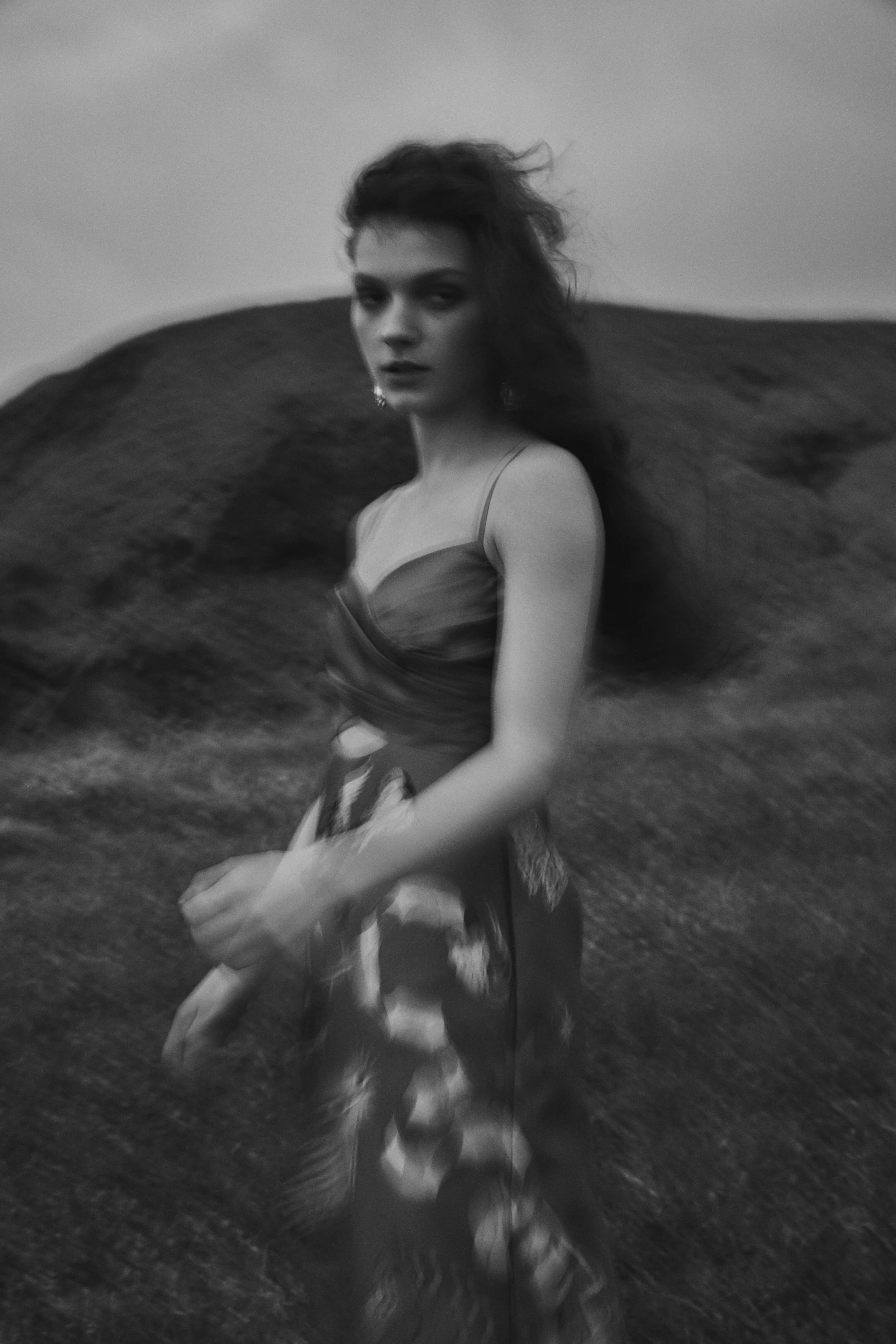 Black and white photo of a woman with long, wavy hair, wearing a sleeveless dress, standing outdoors in front of a large hill or mountain.