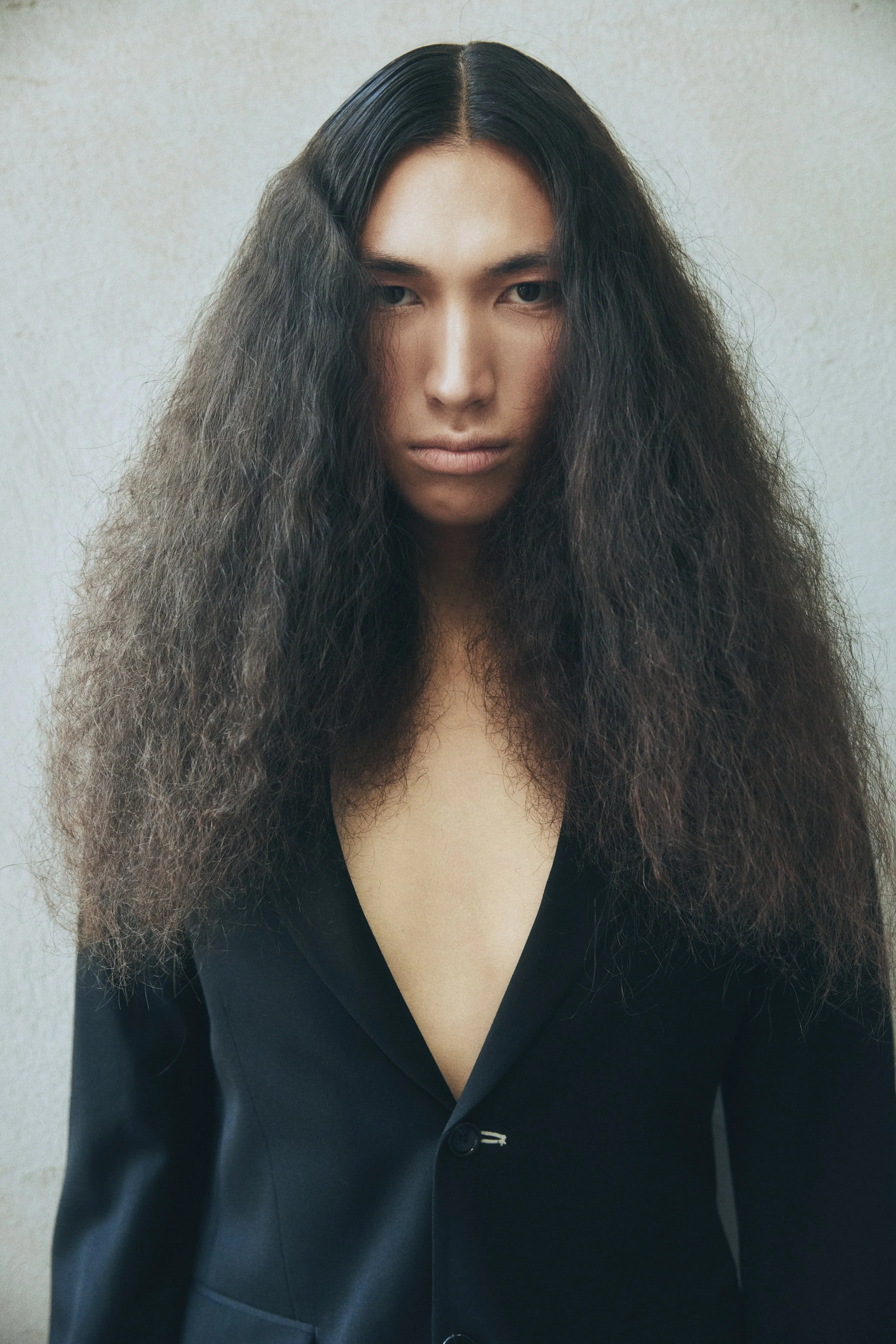 A man with long, curly dark hair wearing a black blazer with a deep V neckline, standing against a plain, light background.