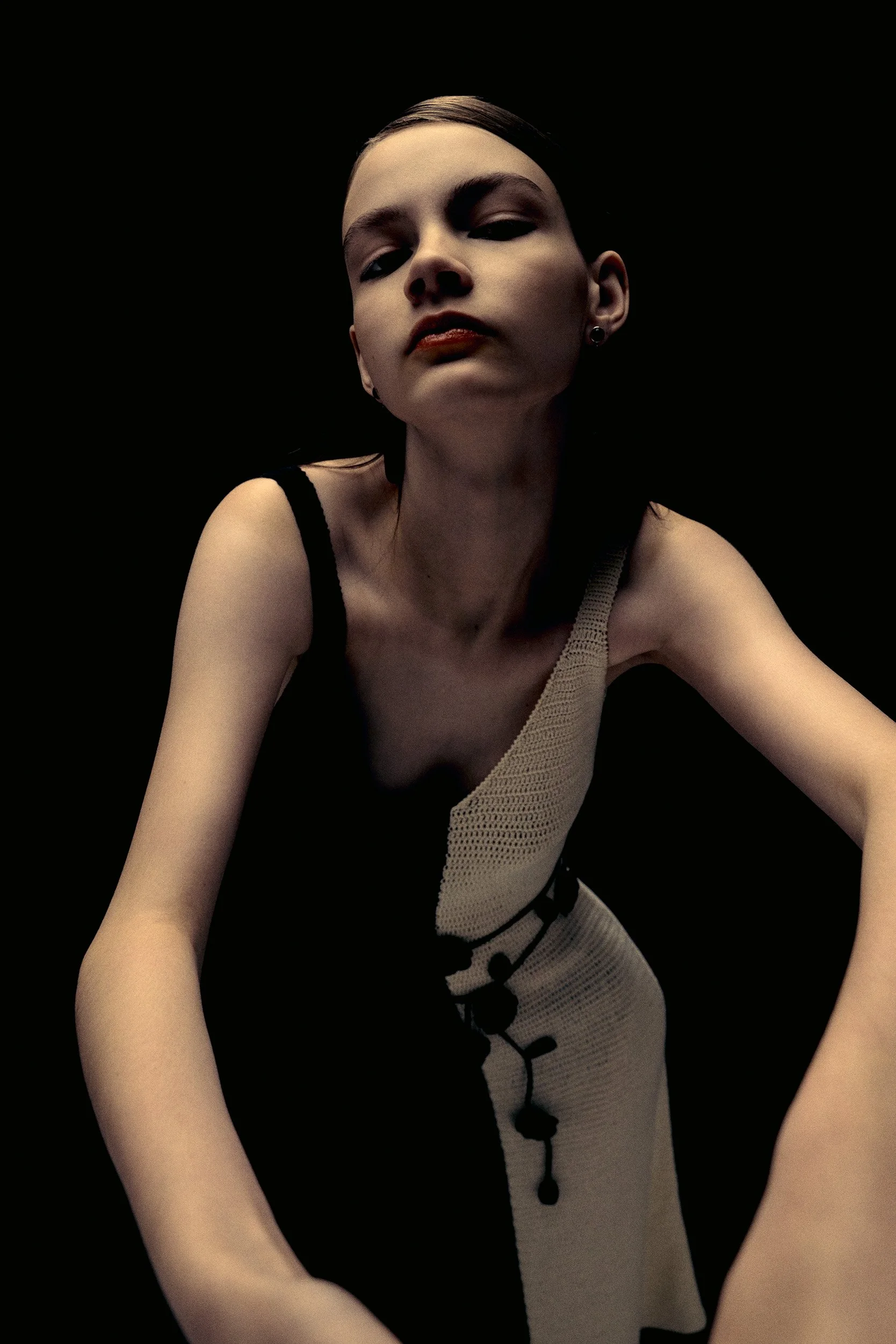 A young woman with short dark hair, fair skin, and bold makeup, wearing a beige and black sleeveless dress, posing against a black background.