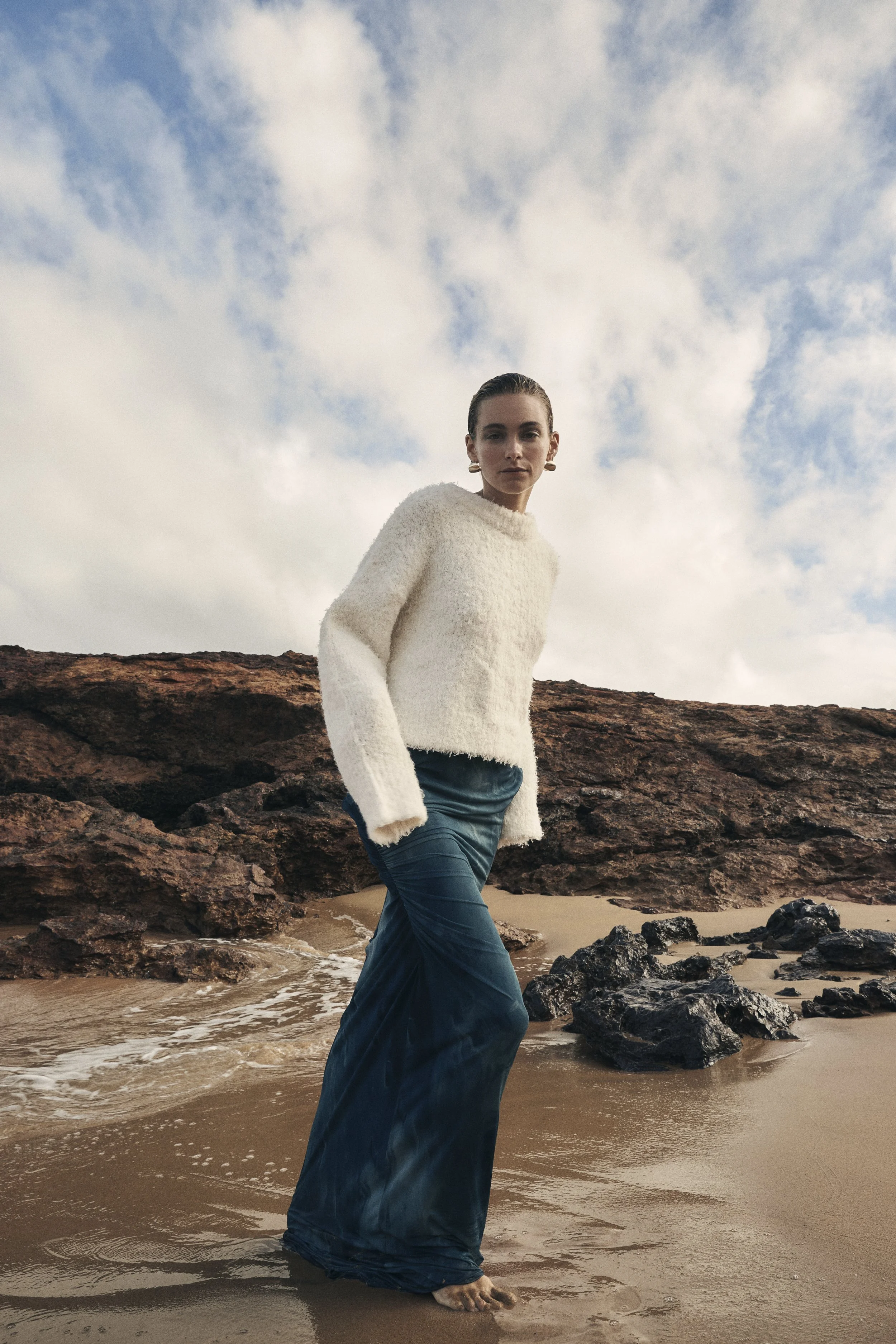 A woman standing on a sandy beach with rocks and a cloudy sky.