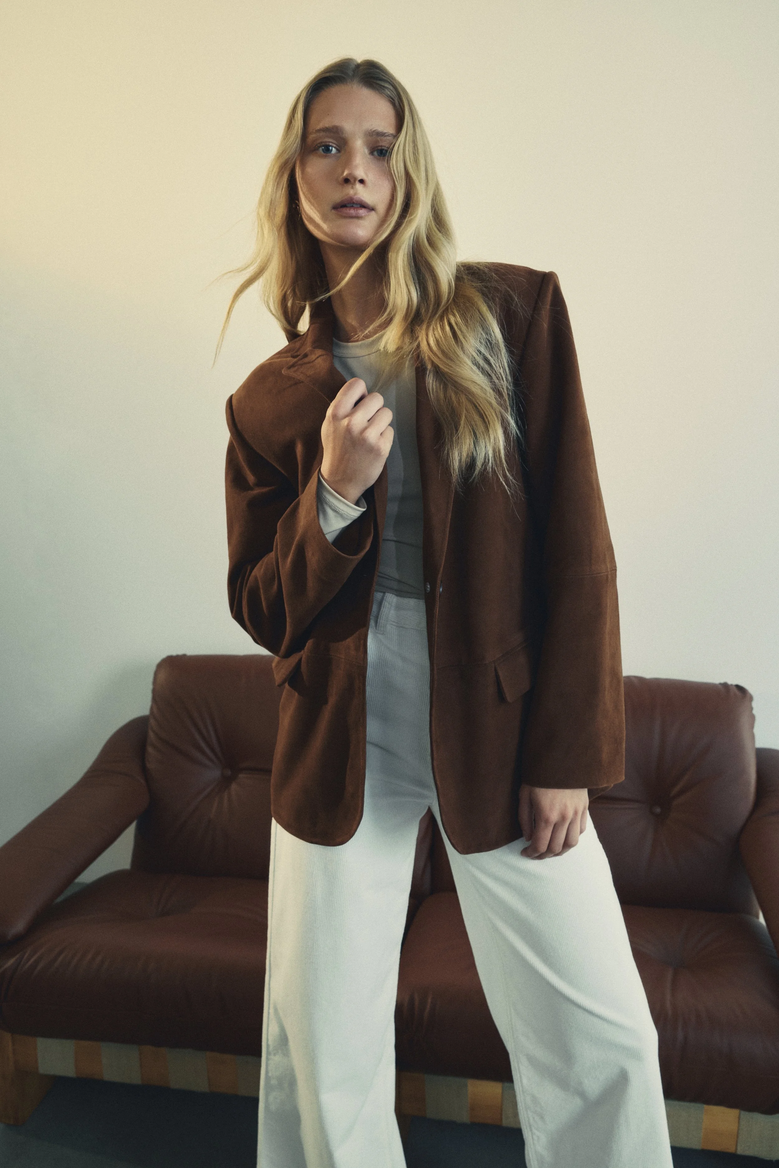 A young woman with long blonde hair wearing a brown blazer over a light-colored top, standing in front of a brown leather couch against a plain wall. Warm light hits the wall behind her.