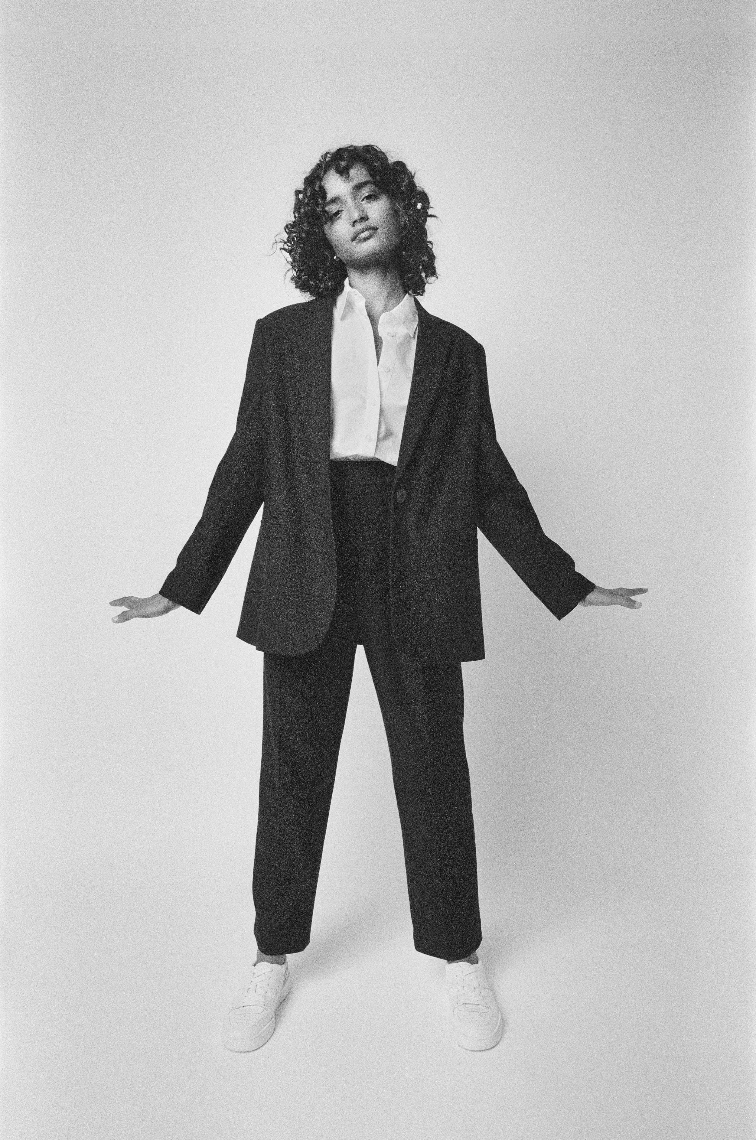 Black and white photo of a woman with curly hair wearing a suit with a blazer, white shirt, and sneakers, standing against a plain background with arms extended outward.