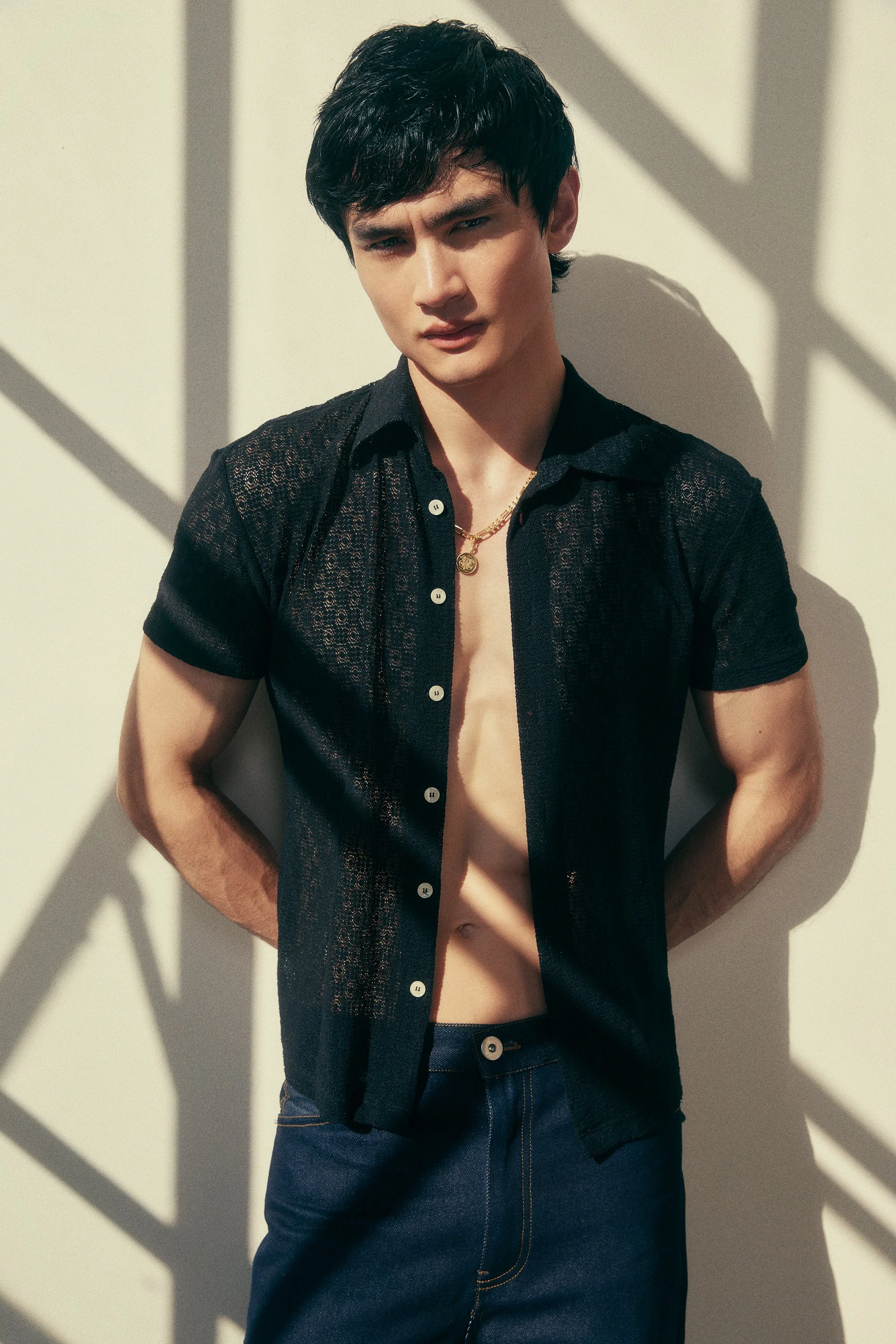 A young man with dark hair and light skin standing against a white wall with shadow patterns. He is wearing an open black, lace-textured shirt, exposing his bare chest and abdomen, along with dark jeans and gold jewelry around his neck.