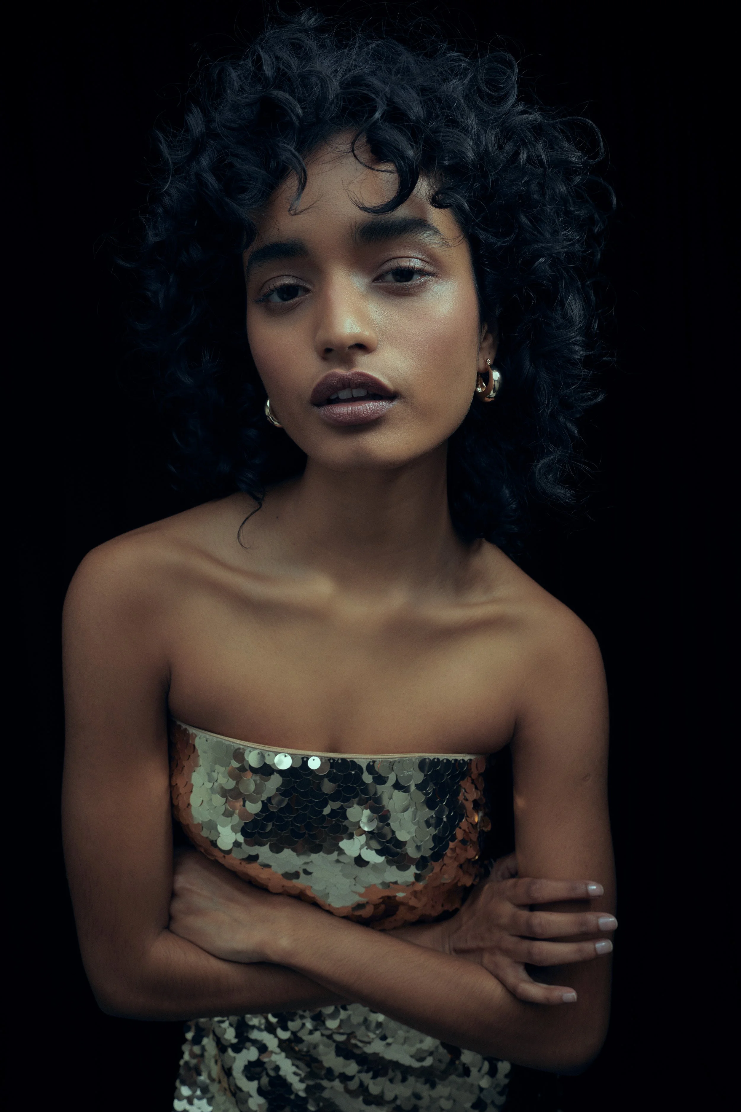 A young woman with curly black hair and hoop earrings, wearing a strapless dress covered in gold and silver sequins, posed against a black background.