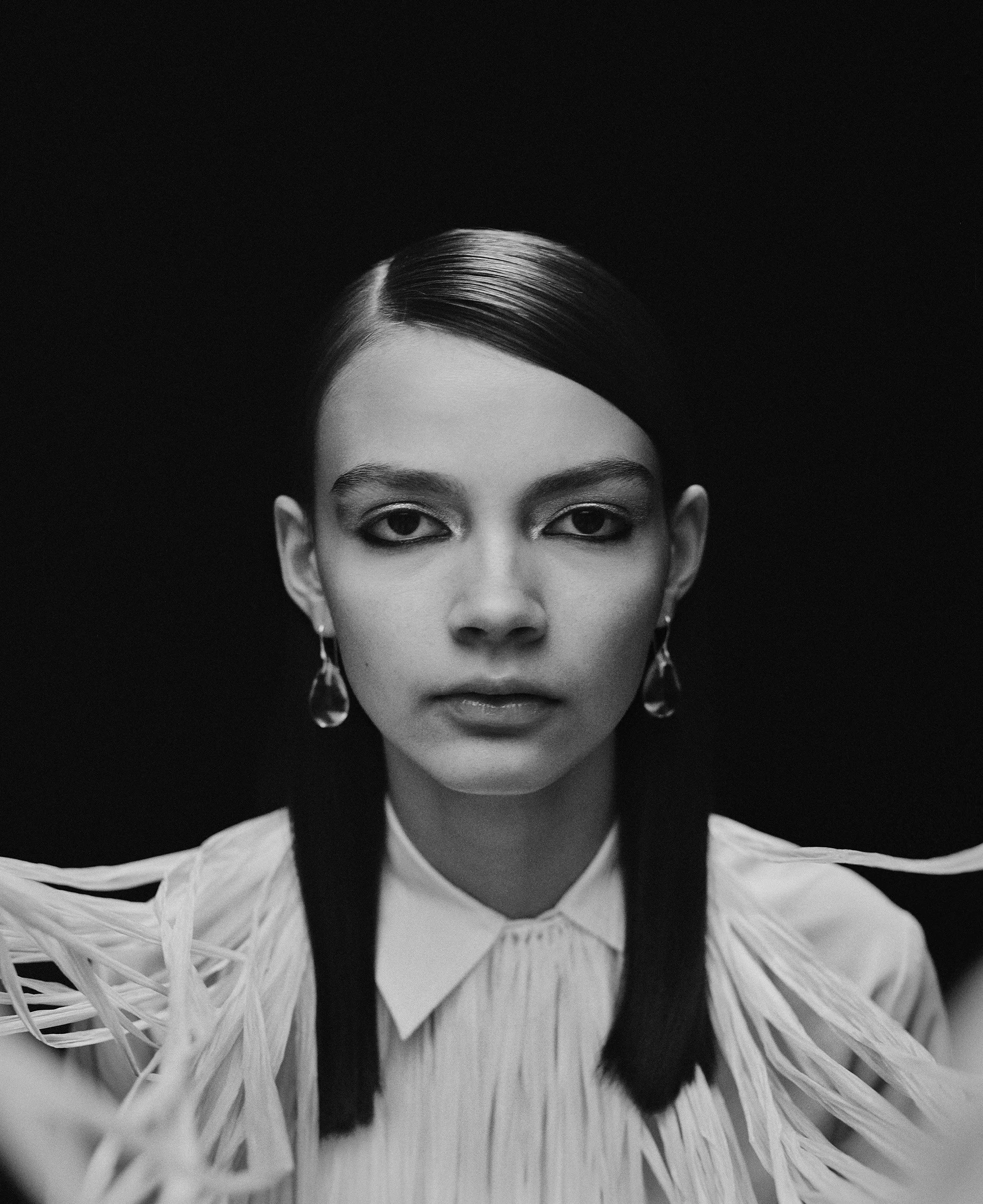 Black and white portrait of a young woman with straight dark hair, wearing earrings and a white shirt with textured detailing, against a dark background.