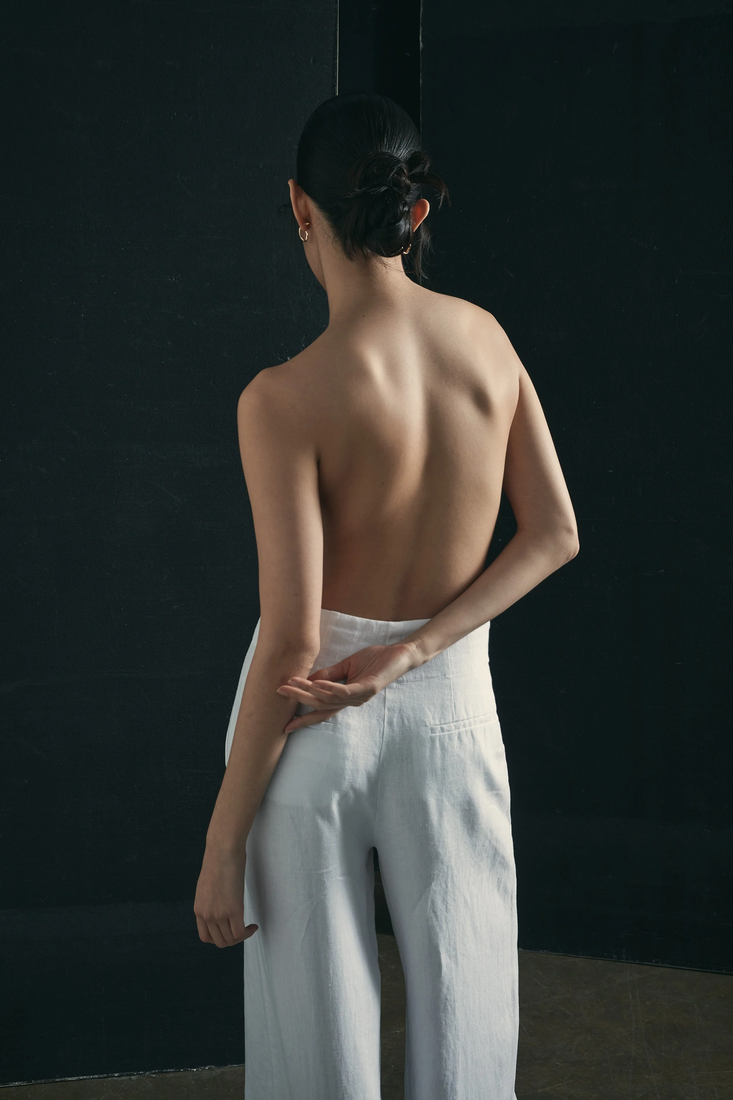 Back view of a topless woman with dark hair in a bun, wearing white pants, standing against a dark wall.