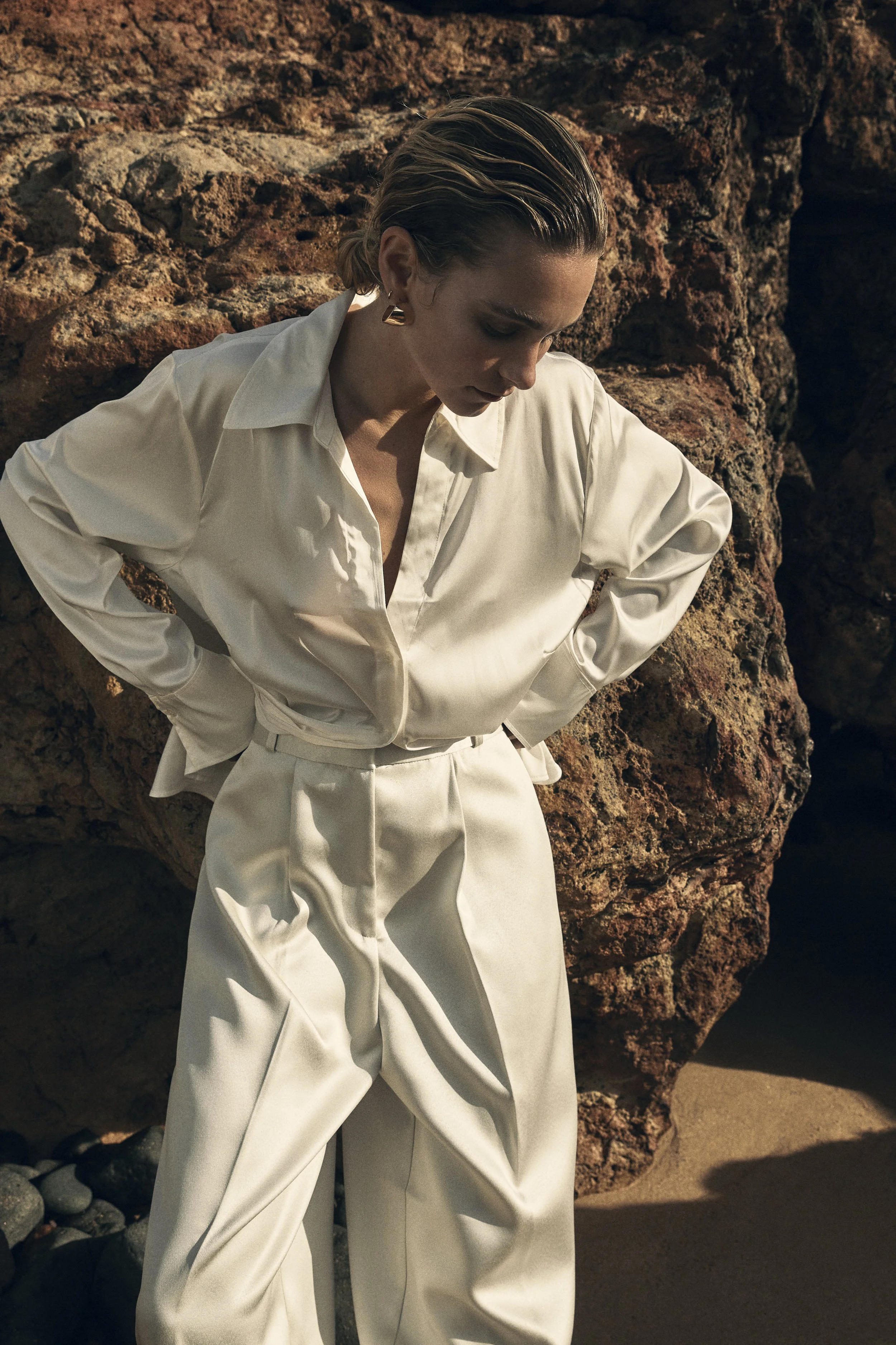 A woman with short blonde hair wearing a cream or off-white satin shirt and matching high-waisted pants, standing with her hands on her hips in front of large reddish-brown rocks on a beach.