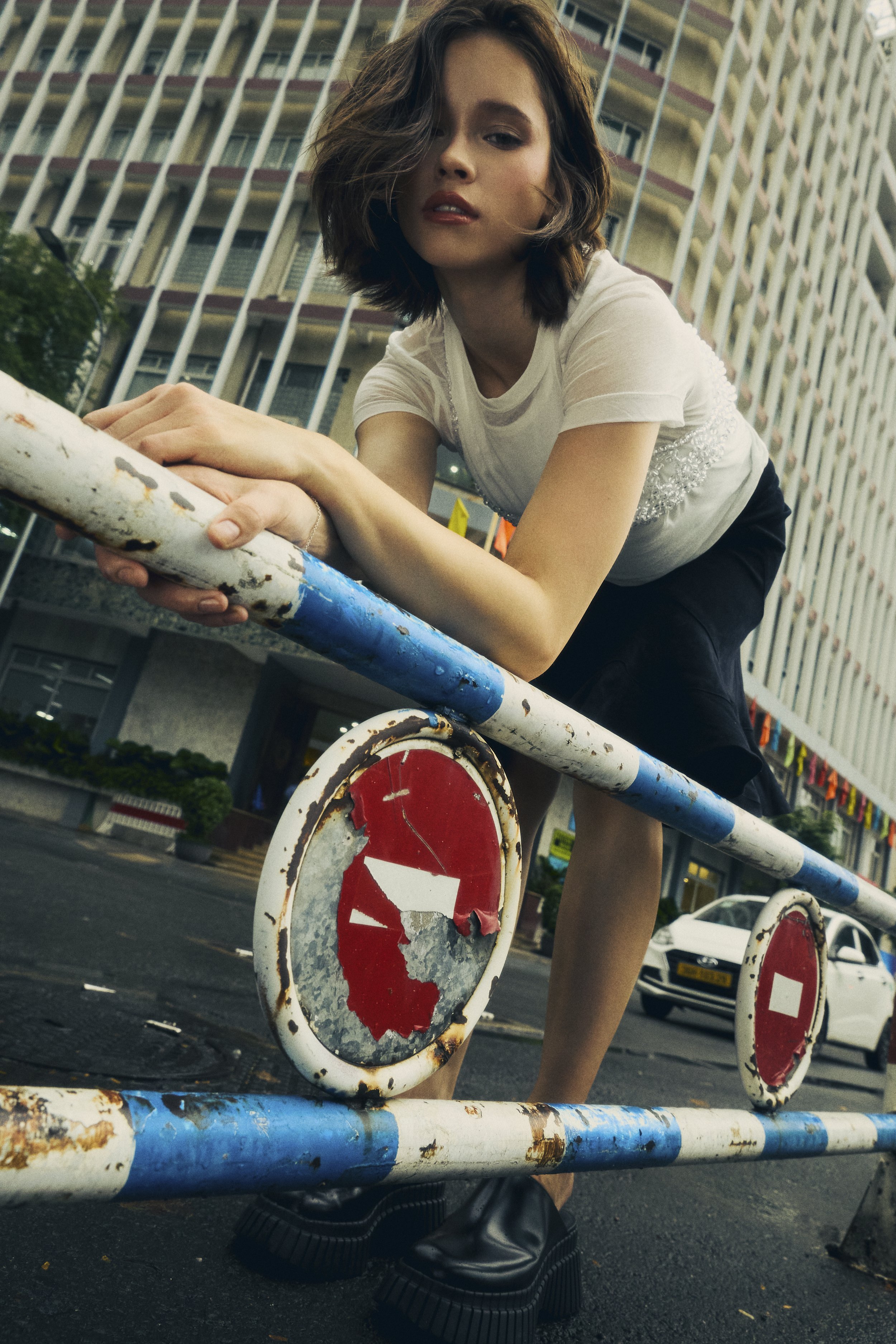 A young woman with short, wavy brown hair leaning on a rusty metal barricade with red and white caution signs, wearing a white T-shirt and black shorts, in an urban setting with tall buildings and a car in the background.
