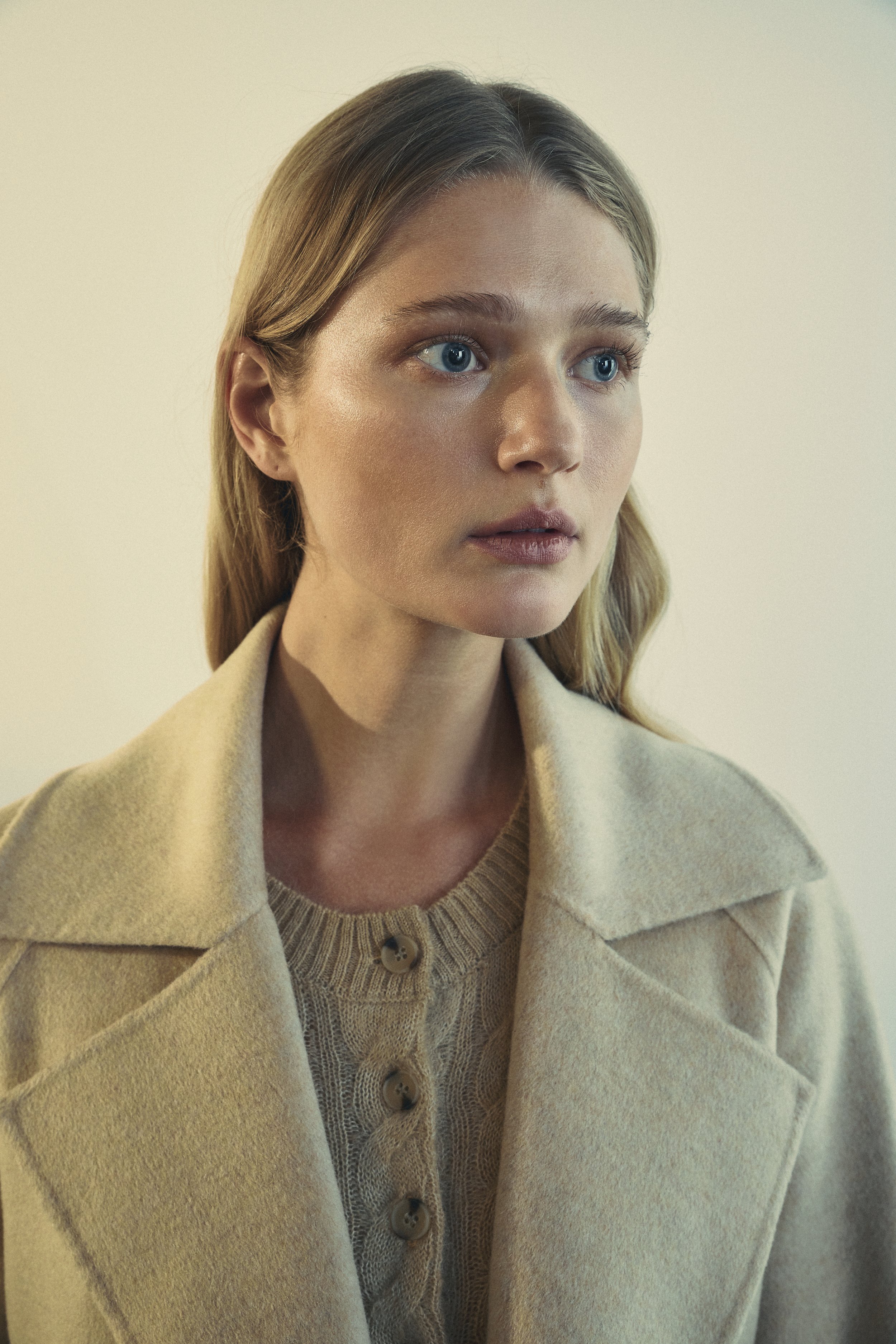 A woman with blonde hair and blue eyes, wearing a beige coat over a light brown knit top with buttons, against a light-coloured sun kissed background.