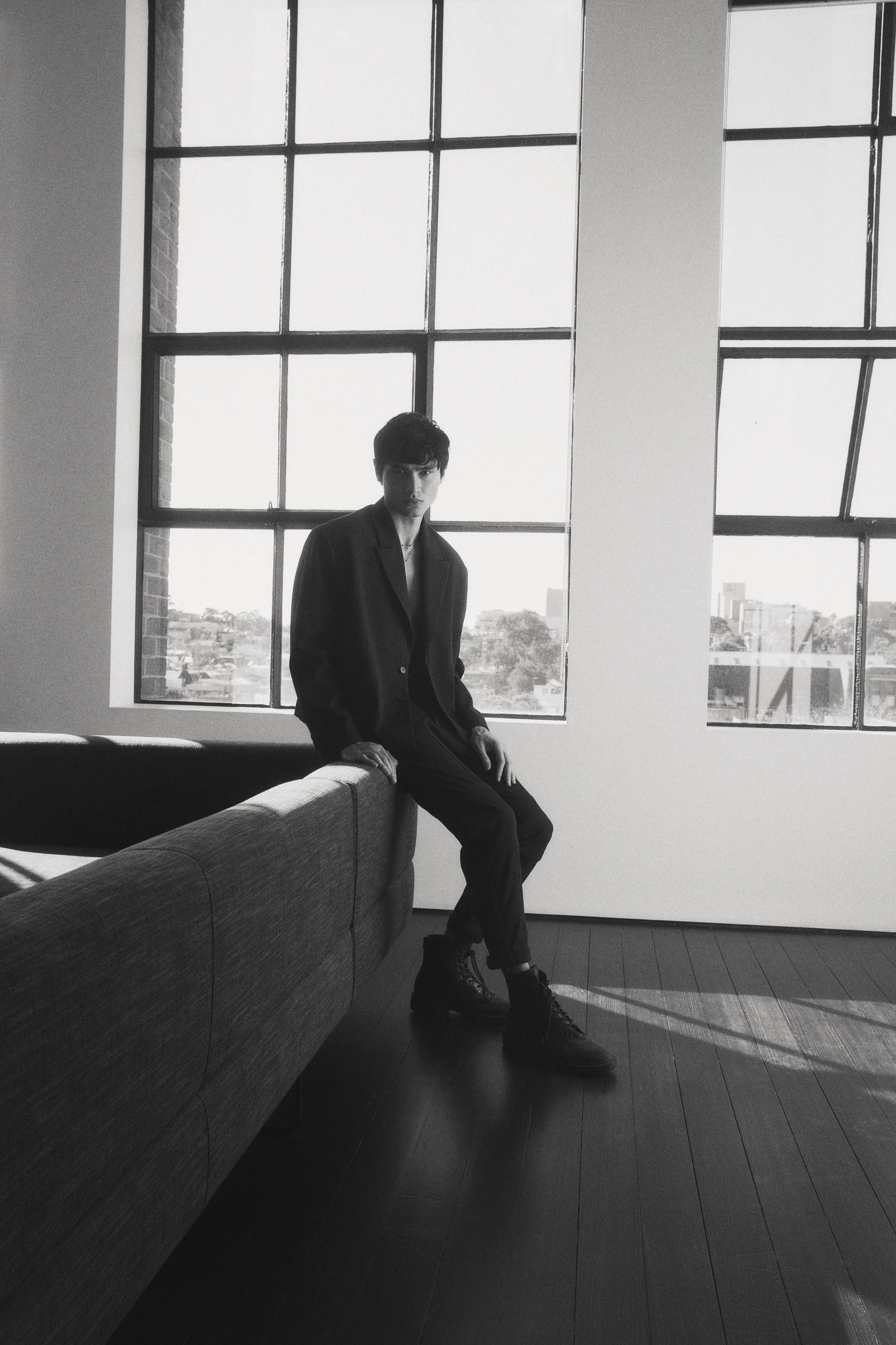 Black and white photo of a young man with dark hair, wearing a dark blazer, sitting on a sofa by large industrial-style windows with a cityscape view in the background, in a modern loft-style interior.