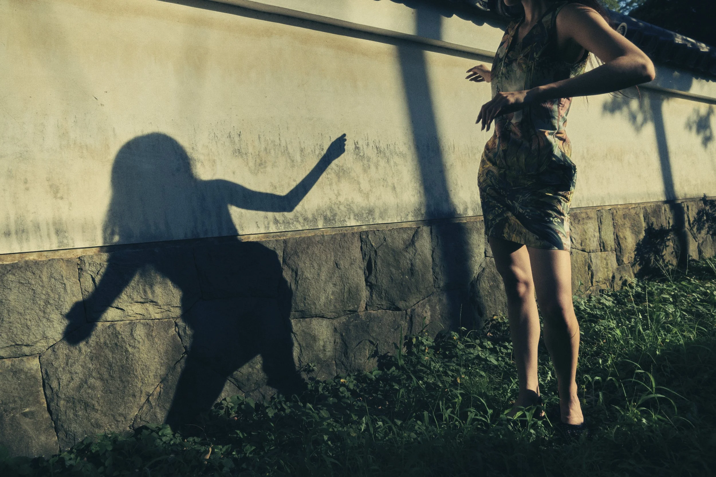 A woman in a patterned dress standing outside near a wall, with her shadow cast on the wall, creating an illusion of a different figure.