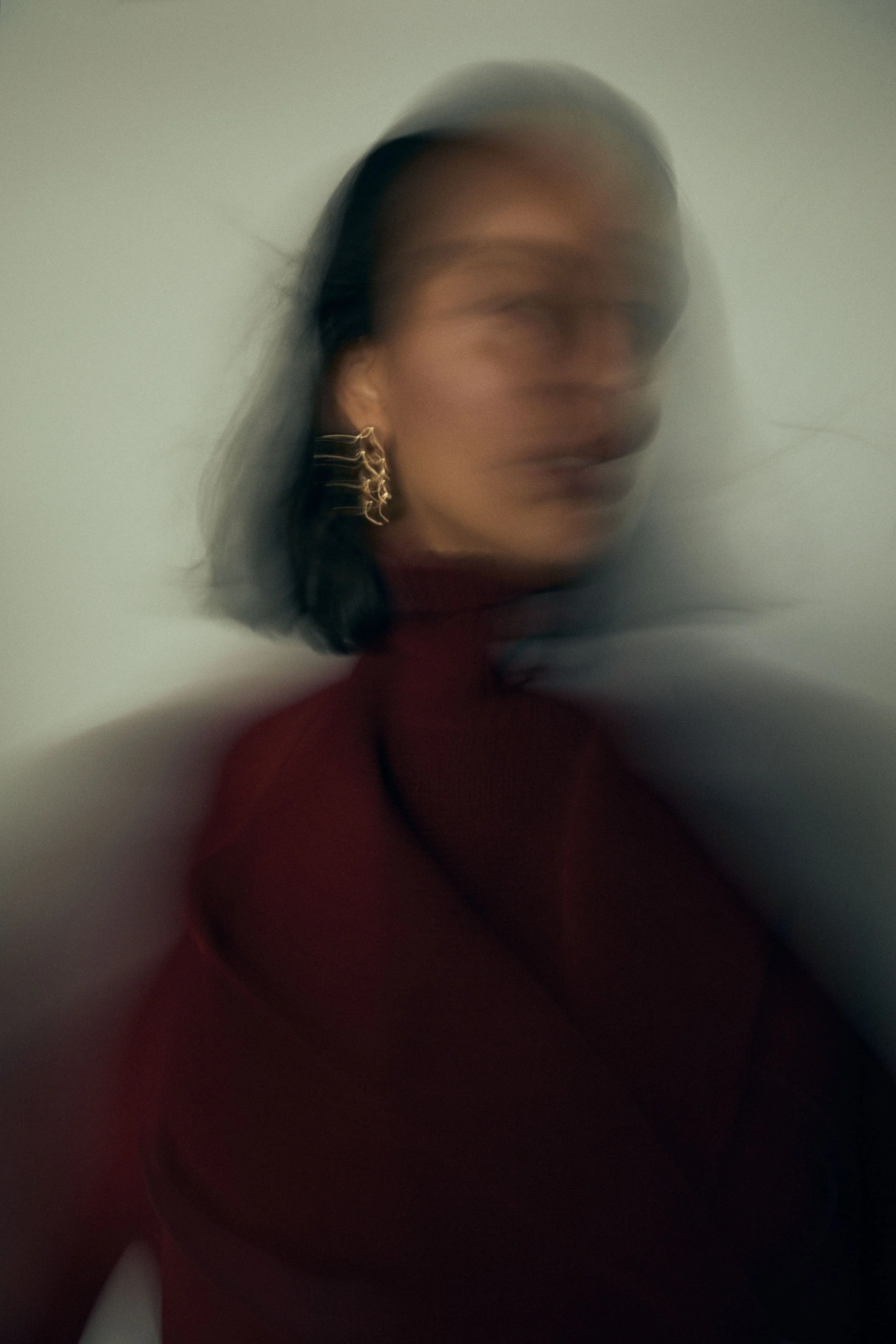 A woman with dark hair wearing a maroon turtleneck and gold earrings, blurred motion effect, standing against a plain light-colored wall.