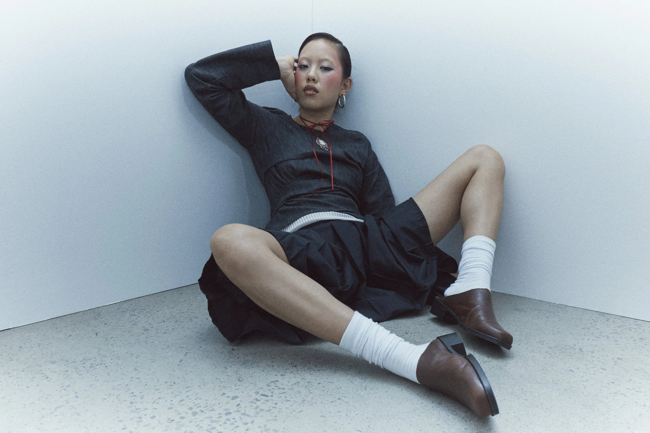 Young woman sitting on the floor against white walls, wearing a dark navy jacket and skirt, white socks, and brown shoes, with her hand resting on her head.