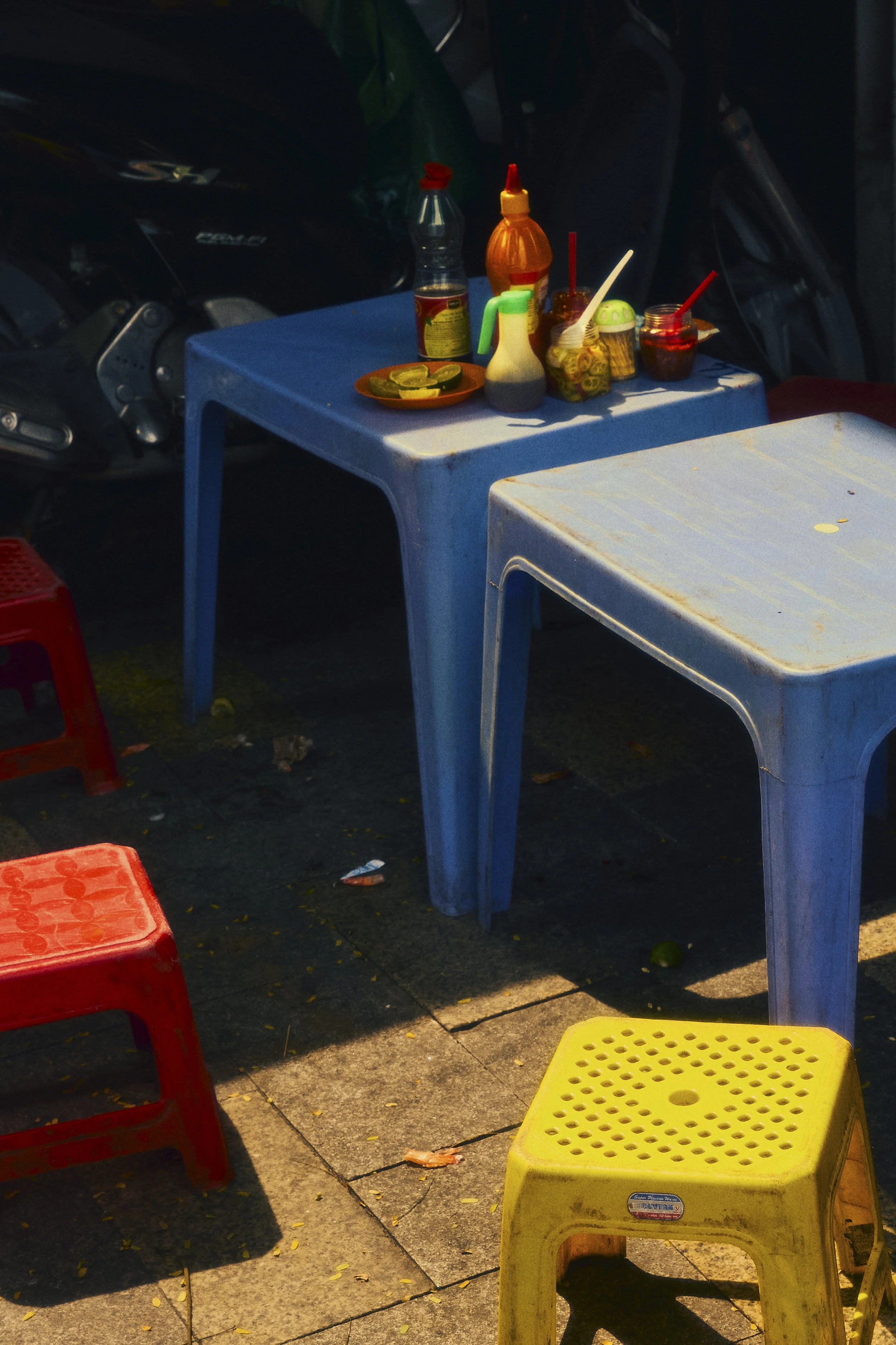 Outdoor scene with a blue plastic table and colorful stools, holding condiments and small dishes, on a sidewalk near parked motorised scooter.