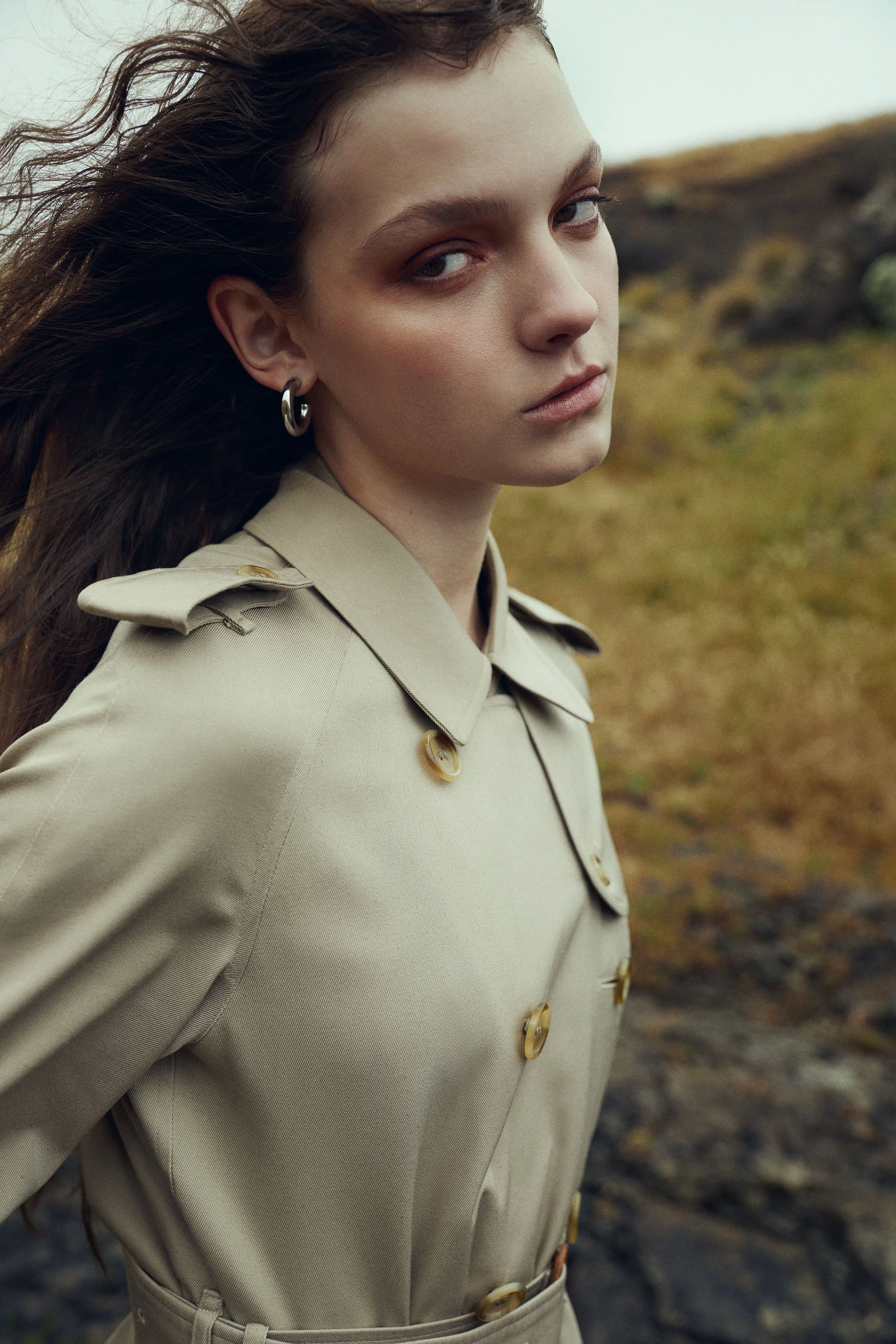 A young woman with long, wavy brown hair standing outdoors in a natural landscape, wearing a beige trench coat with epaulets and large buttons, looking serious with a neutral expression.