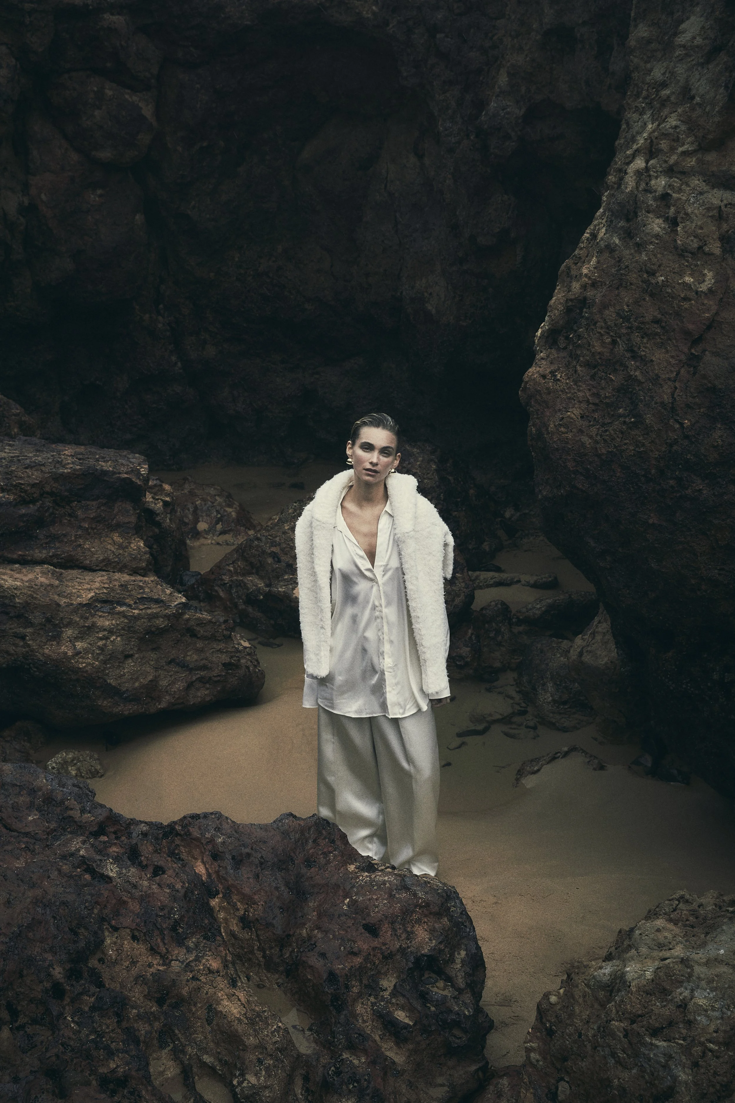 A woman in white silk clothing and a white jacket stands on a sandy beach surrounded by large rocks and dark caves.