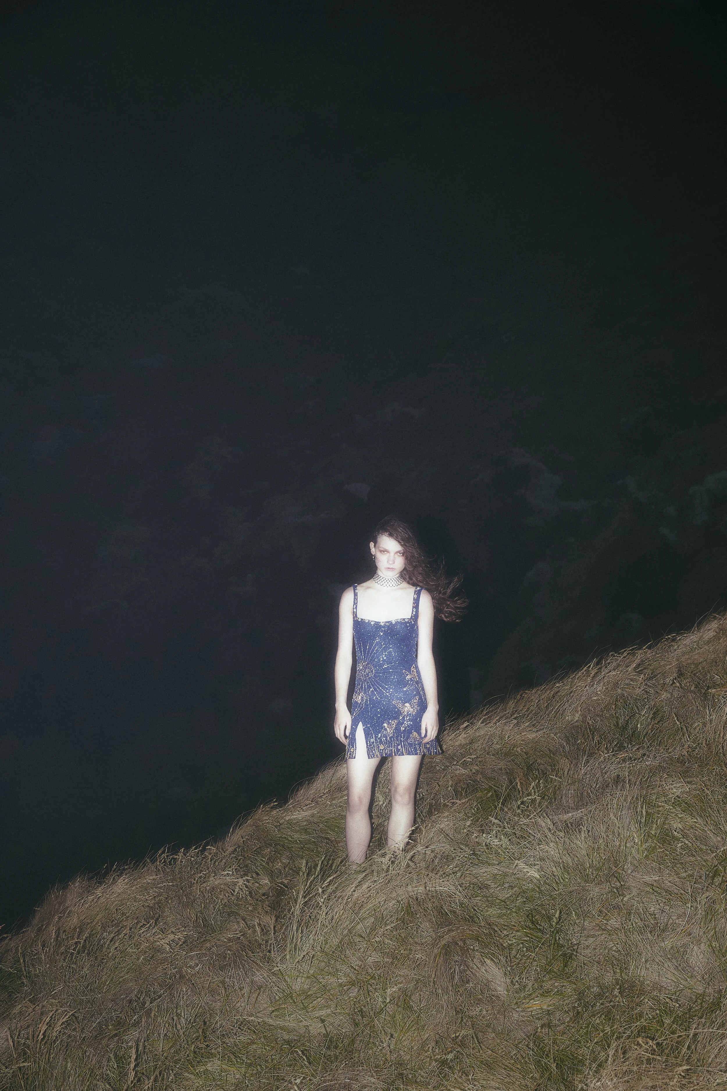 A woman in a blue dress standing on a grassy hill at night with a dark cloudy sky