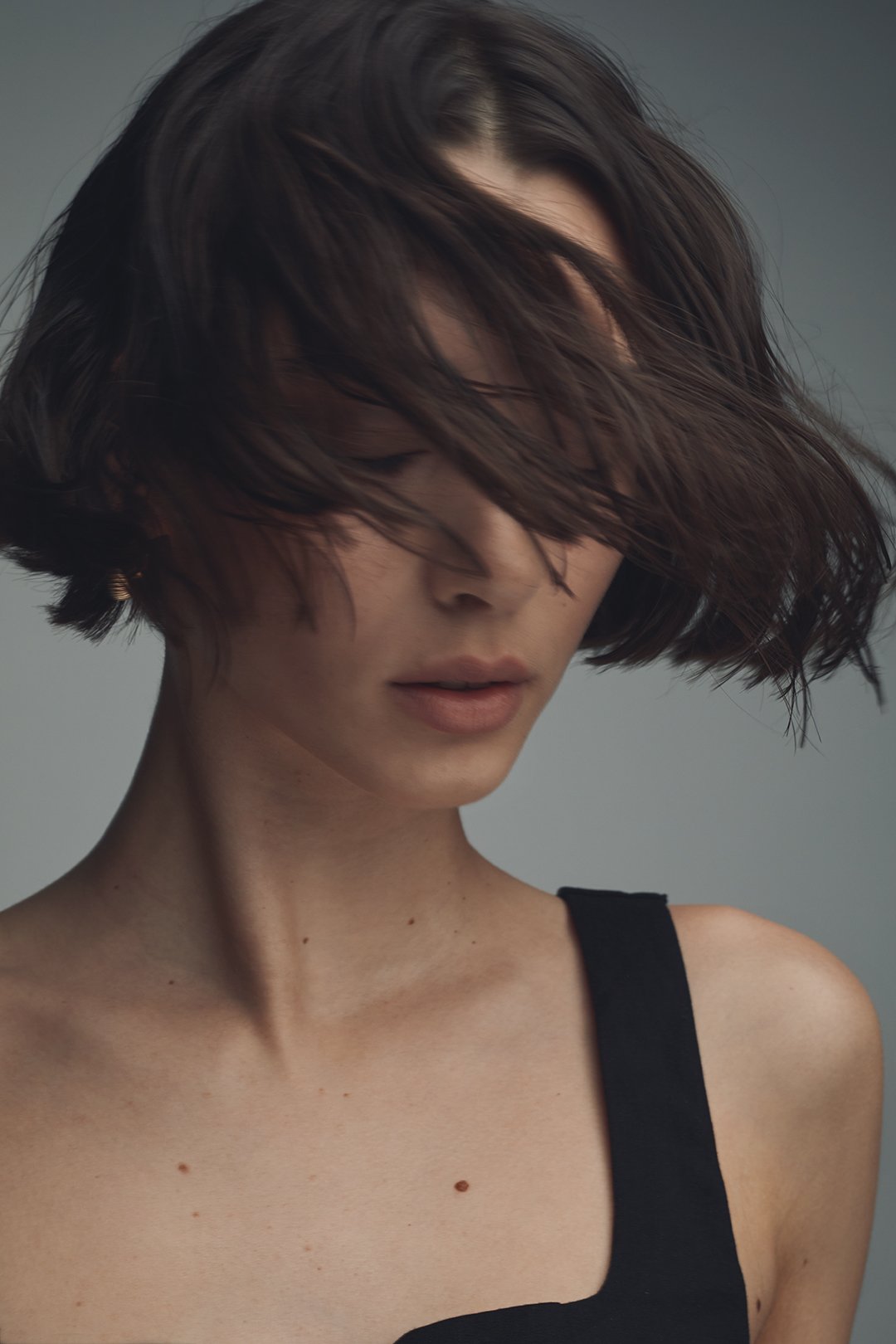 A slightly motion blurred woman with short, dark, and wavy hair covering part of her face, wearing a black sleeveless top, against a neutral grey background.