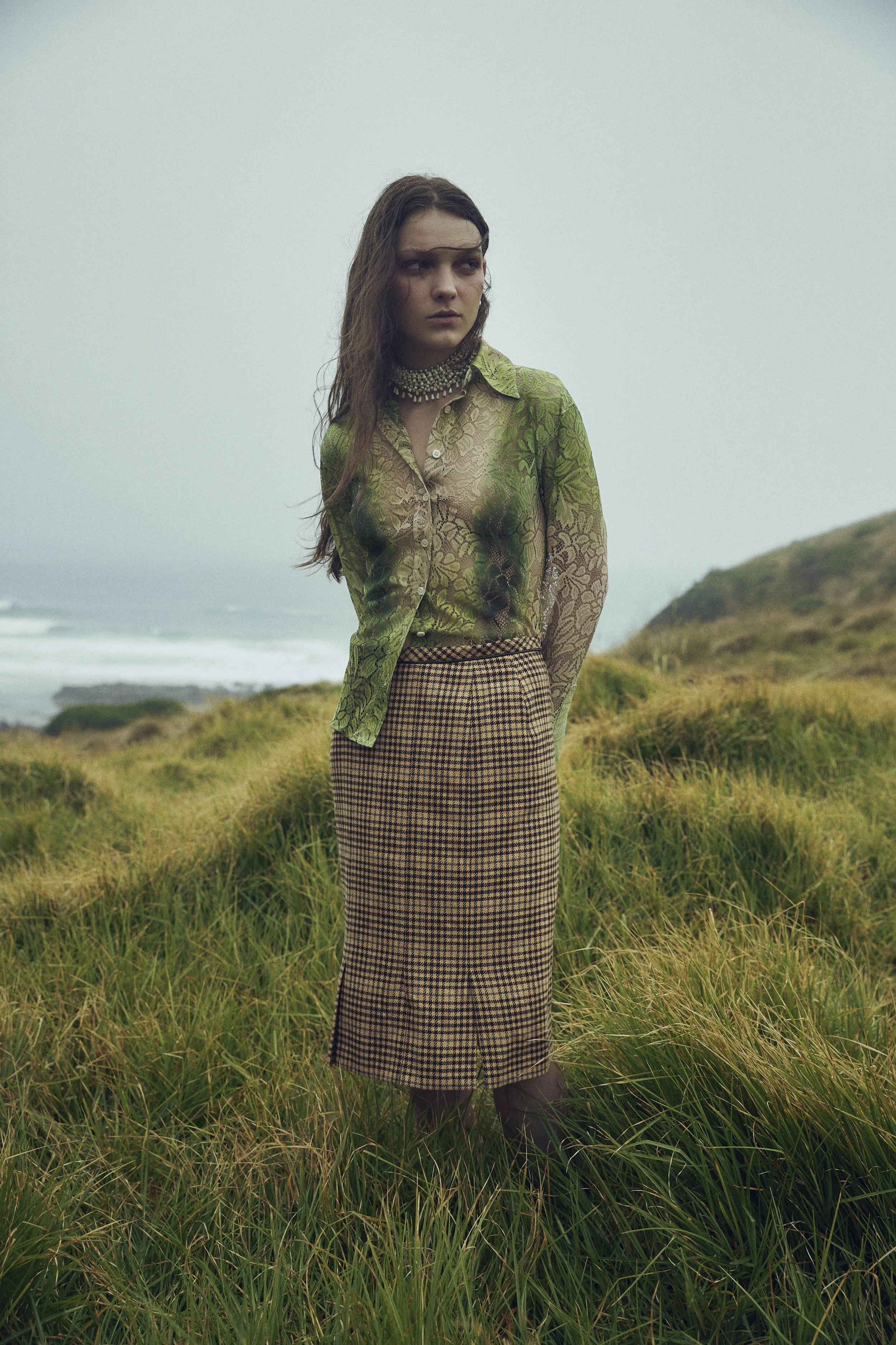 A woman with long hair stands outdoors in a grassy area with hills and the ocean in the background, wearing a sheer green lace blouse, a chunky necklace, and a plaid skirt.