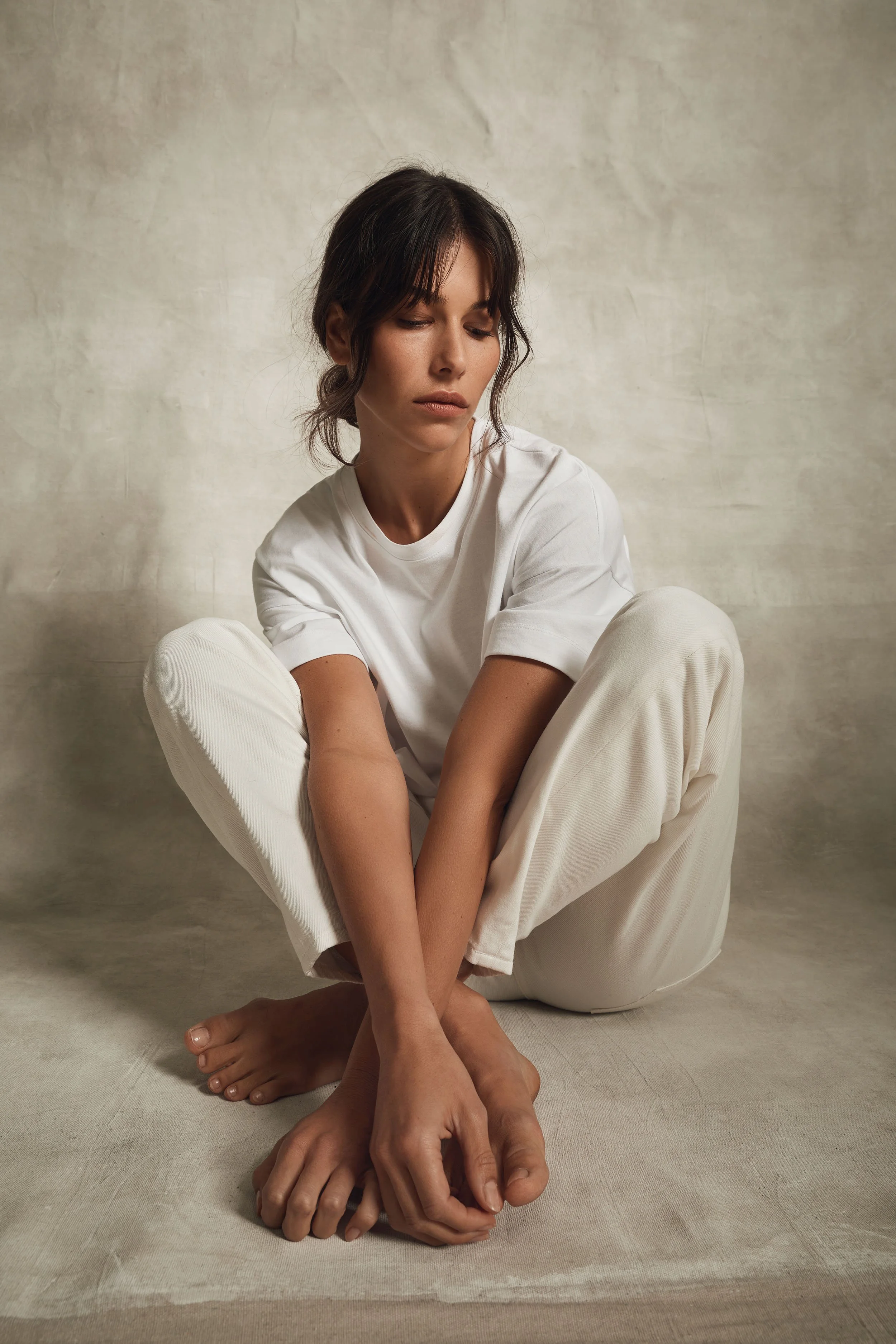 A woman with dark hair sitting on the floor with her knees drawn to her chest, wearing a white t-shirt and cream-colored pants, against a textured beige wall.