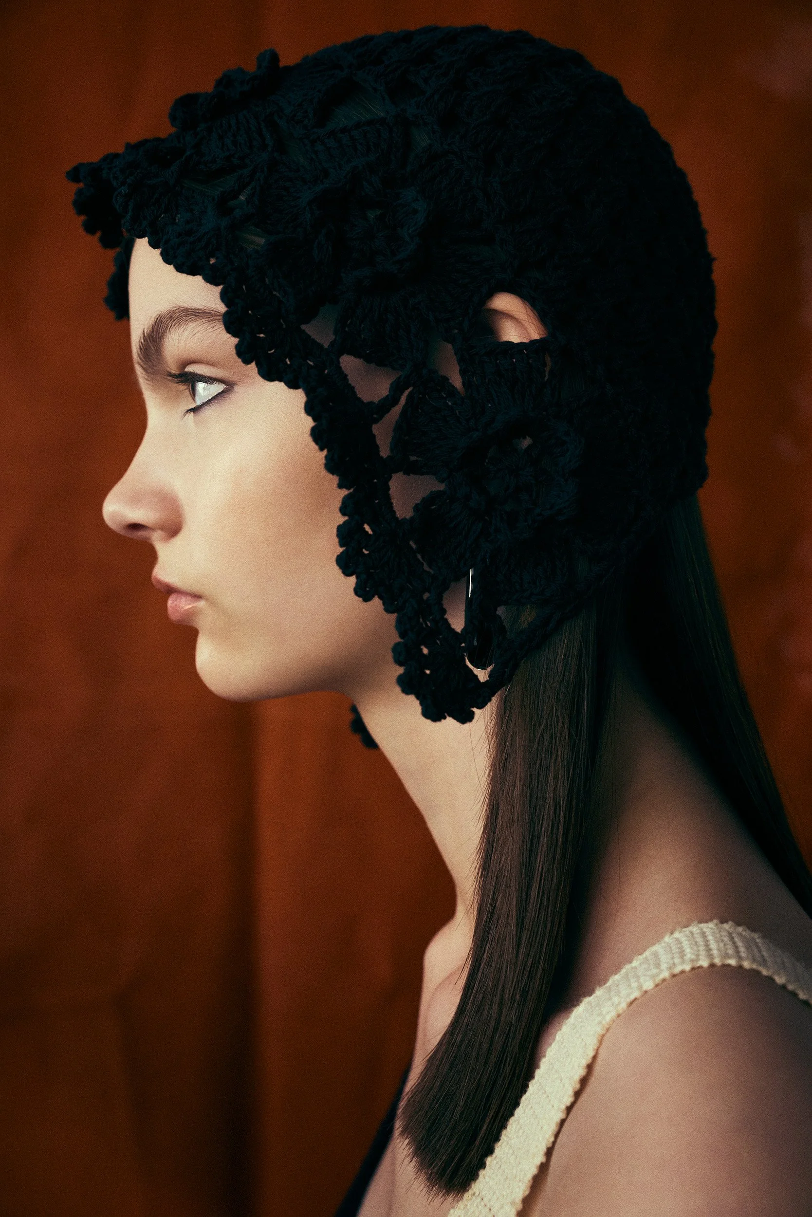 A side profile of a woman with long straight brown hair, wearing a black crocheted headpiece. She has light skin, defined eyebrows, and is wearing a beige strap top. The background is a dark orange colour.