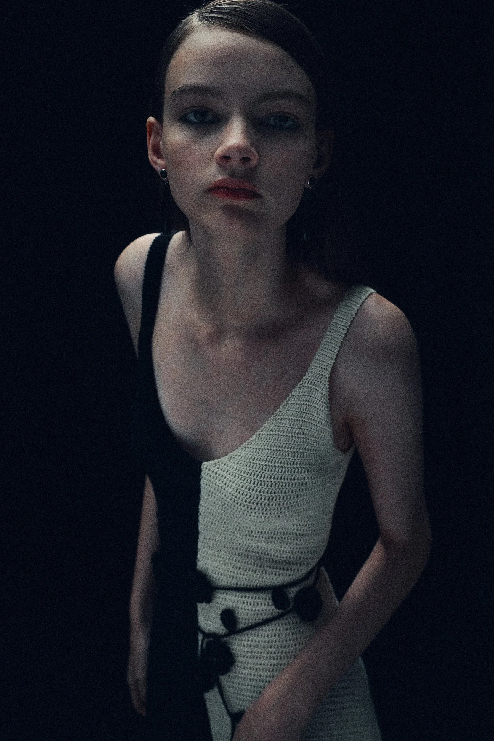 A young woman with dark hair and light skin looking directly at the camera, wearing a sleeveless dress with a black and white pattern, against a dark background.
