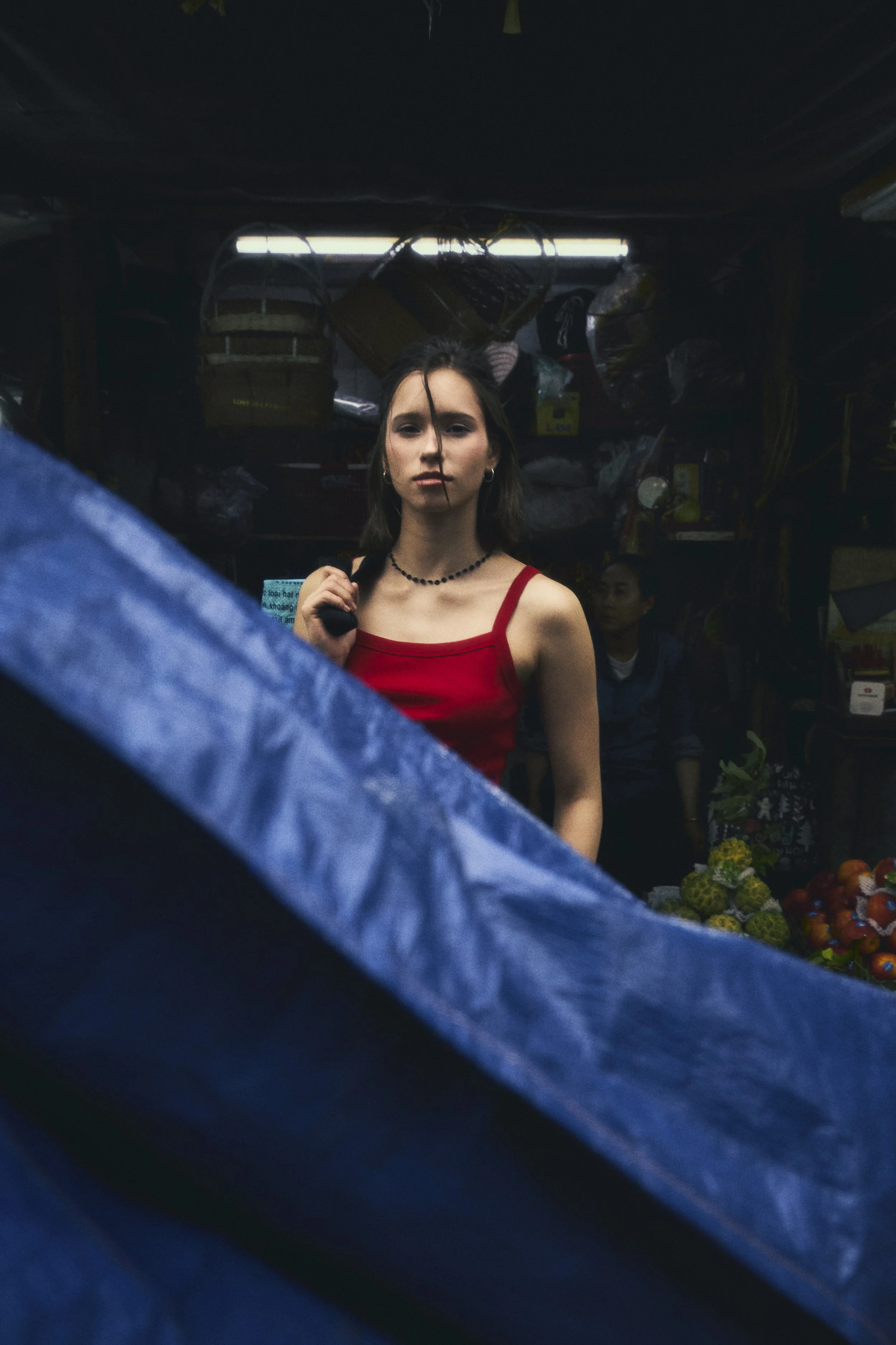 A woman in a red tank top standing in a market, with a serious expression, dark background, fruit visible in the stall.