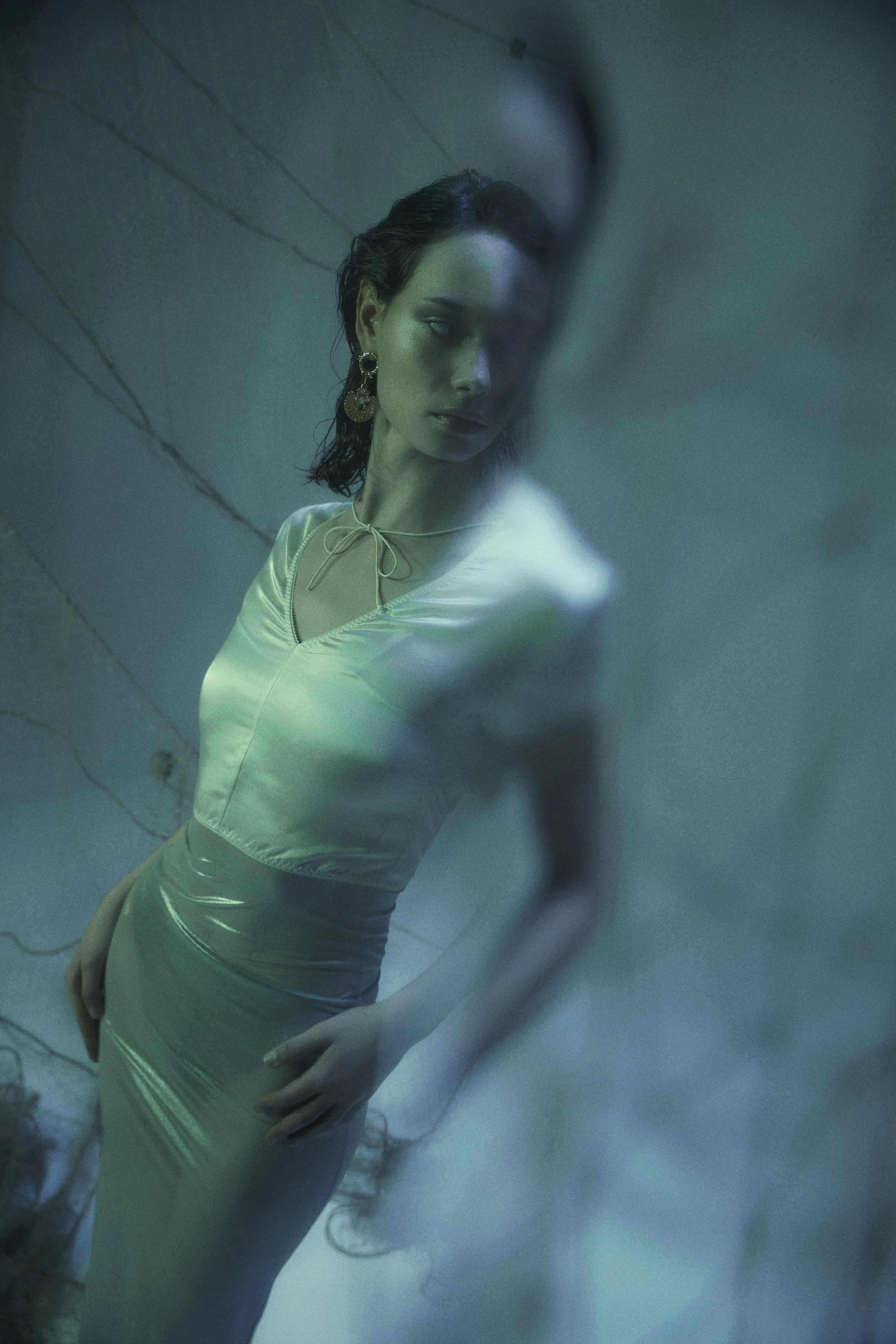 A woman with dark, wet hair and statement earrings, wearing a shiny, satin dress with a front tie detail, standing behind an opaque surface, creating a blurred and surreal visual effect.