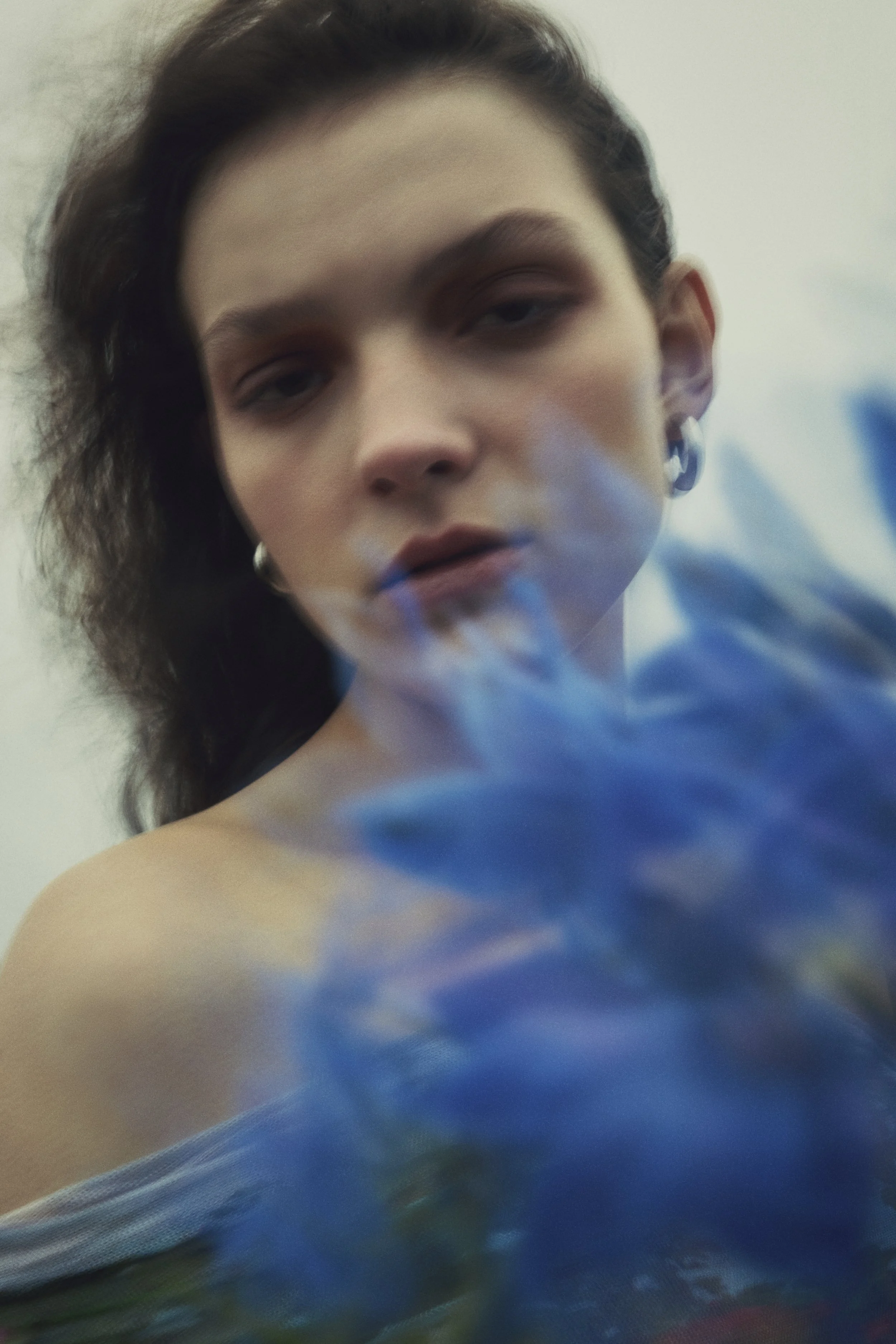 A close-up portrait of a woman with dark, wavy hair and earrings, looking directly at the camera with slightly parted lips. She is partially obscured by out-of-focus blue flowers in the foreground.