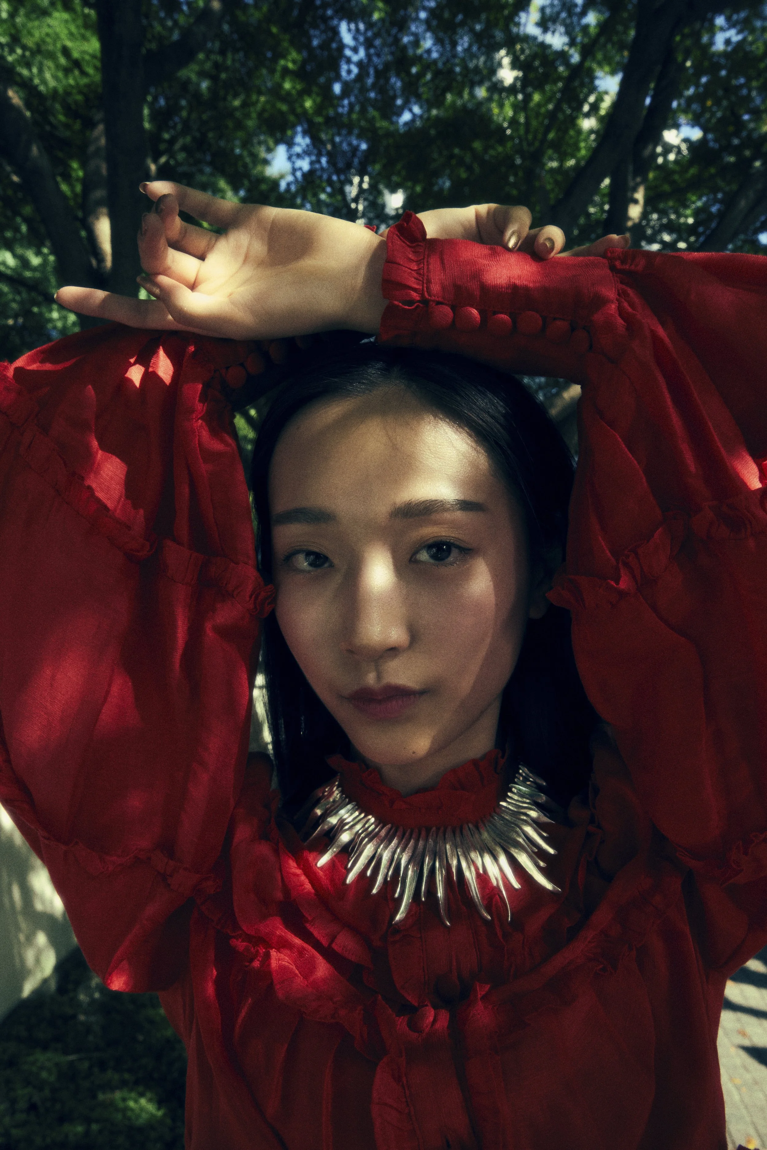 A woman in a red dress with long dark hair, standing outdoors under trees with sunlight filtering through, looking directly at the camera with a neutral expression. She is wearing a statement necklace with metallic spikes.