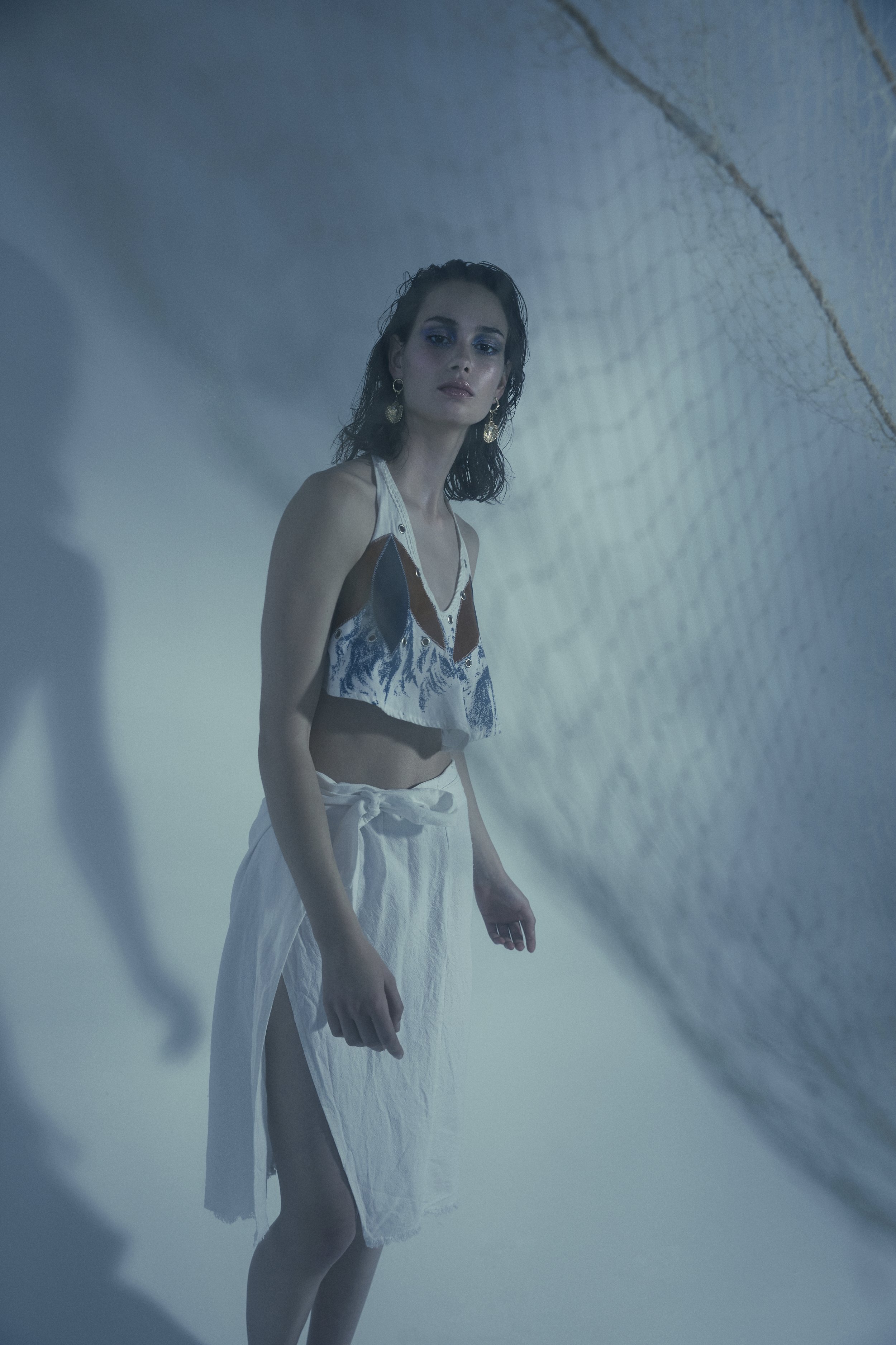 A young woman with dark, wet hair and earrings stands in a blue-lit, textured setting wearing a white, cut-out top with a blue and brown design and a white skirt.