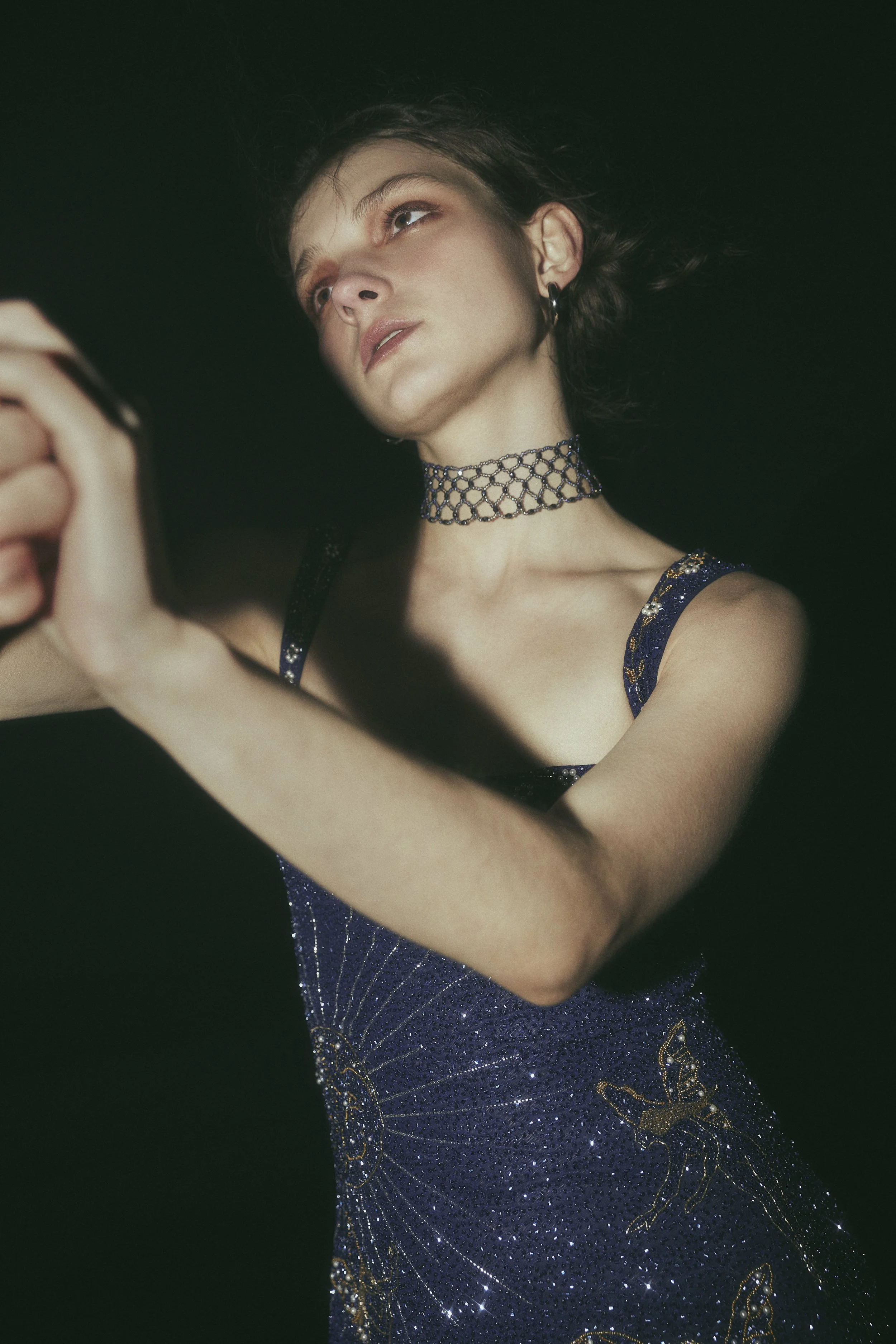 A woman with short brown hair wearing a sparkly navy blue dress with celestial embroidery, a black choker, and hoop earrings, is posing against a dark night sky background.