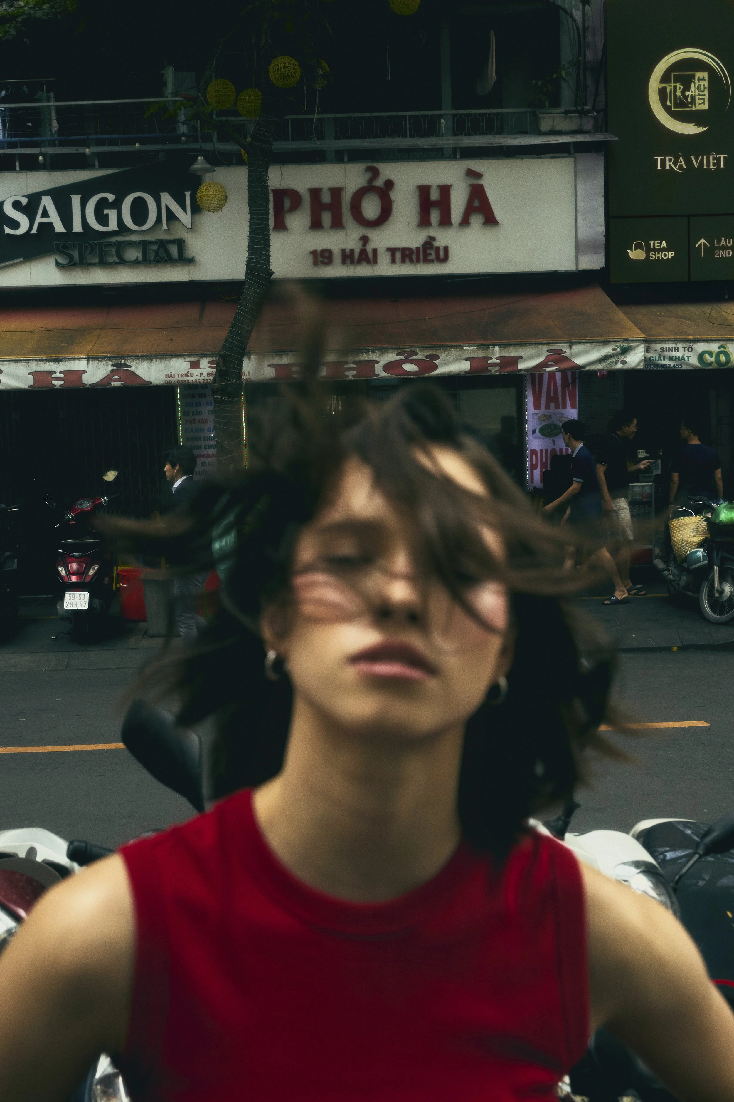 A young woman, out of focus with short hair wearing a red sleeveless top standing in front of an in focus street scene with shop signs, motorcycles, and pedestrians, with her hair being blown by the wind.