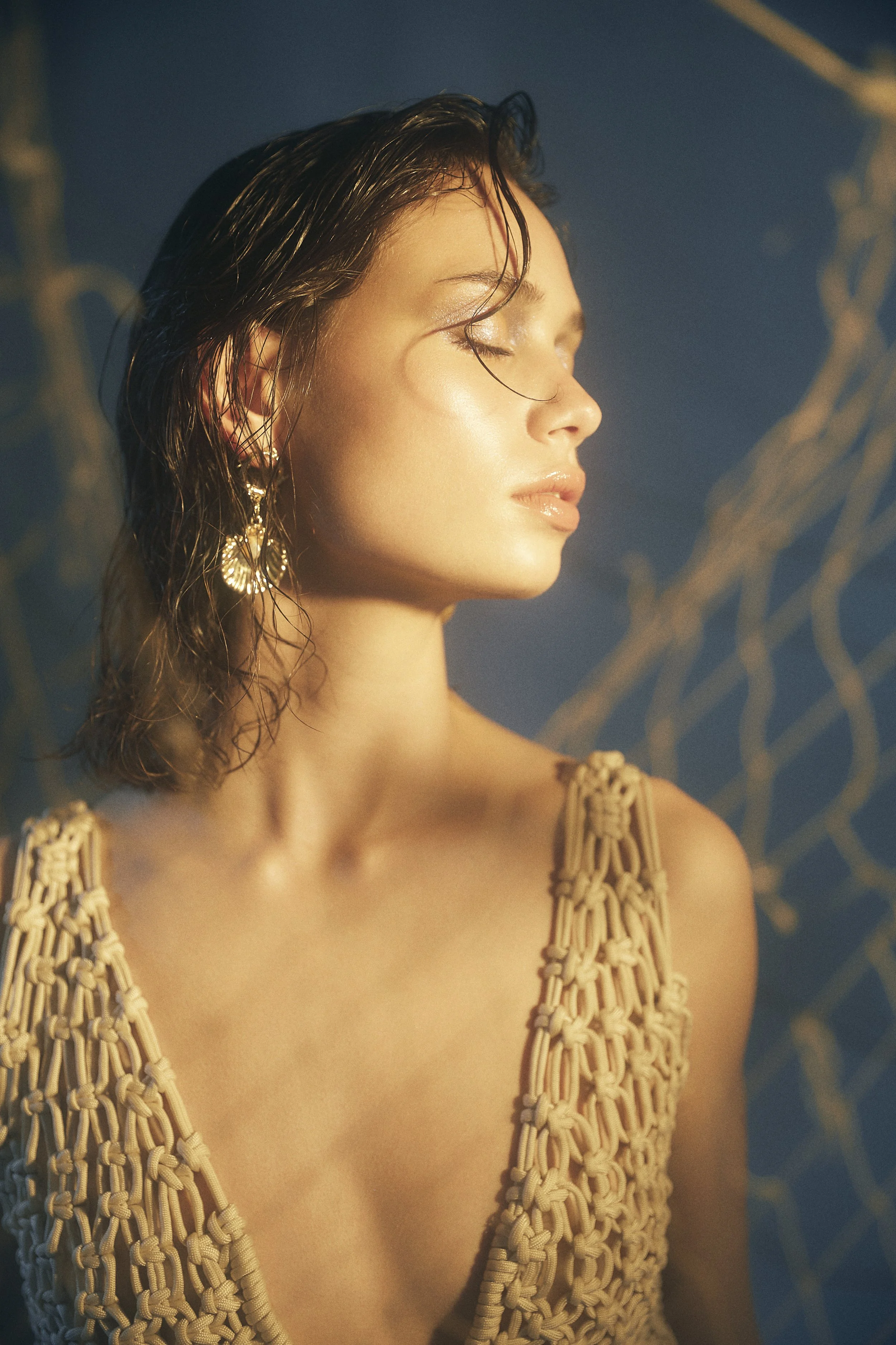 A woman with wet hair and earrings, wearing a macrame top, with her eyes closed, basking in golden light against a blue background with abstract lines.