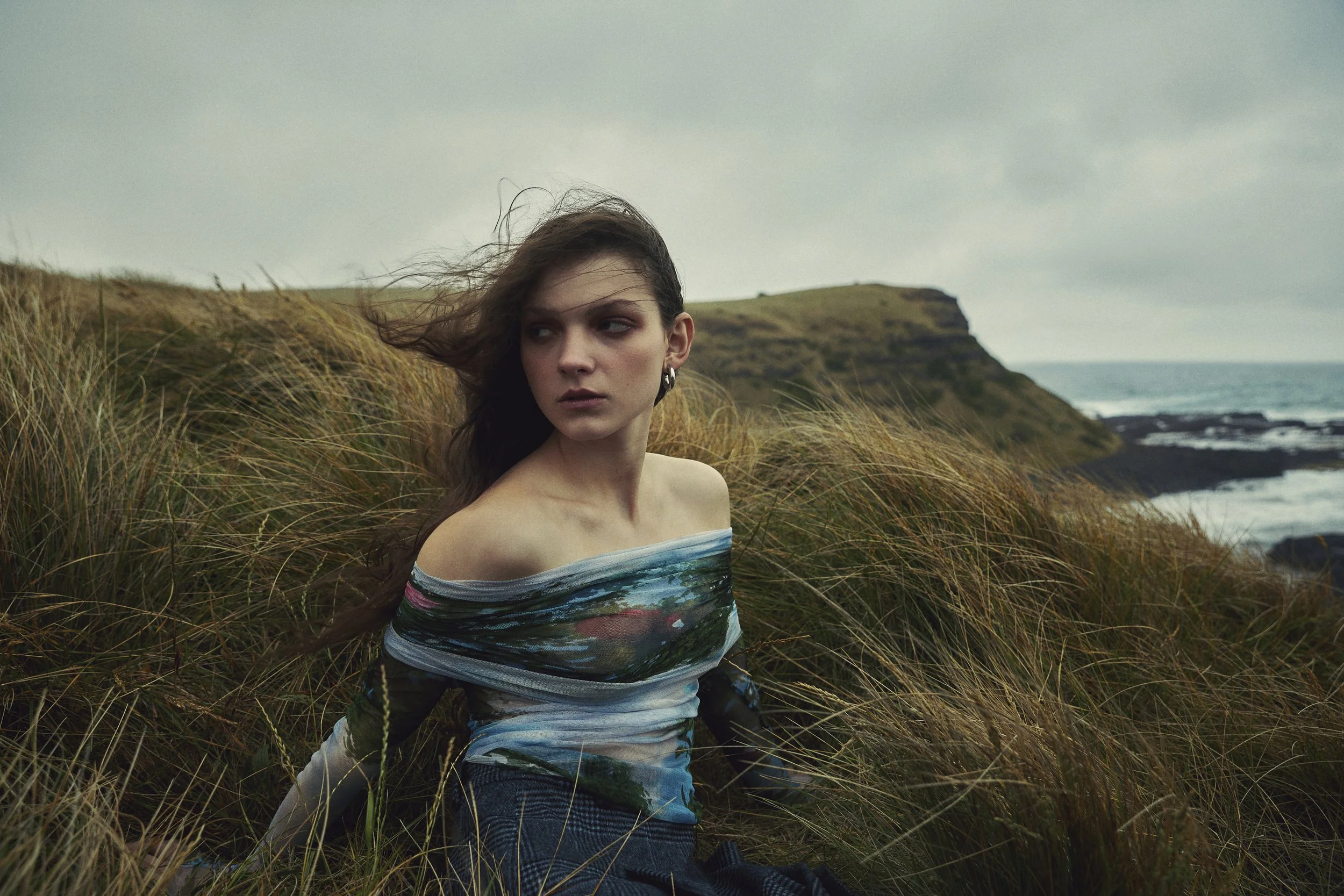 A woman sitting among tall grass on a cloudy day with a cliff and ocean in the background wearing a slightly sheer long sleeve off the shoulder top with an abstract painterly pattern. Her hair blown by the coastal breeze.