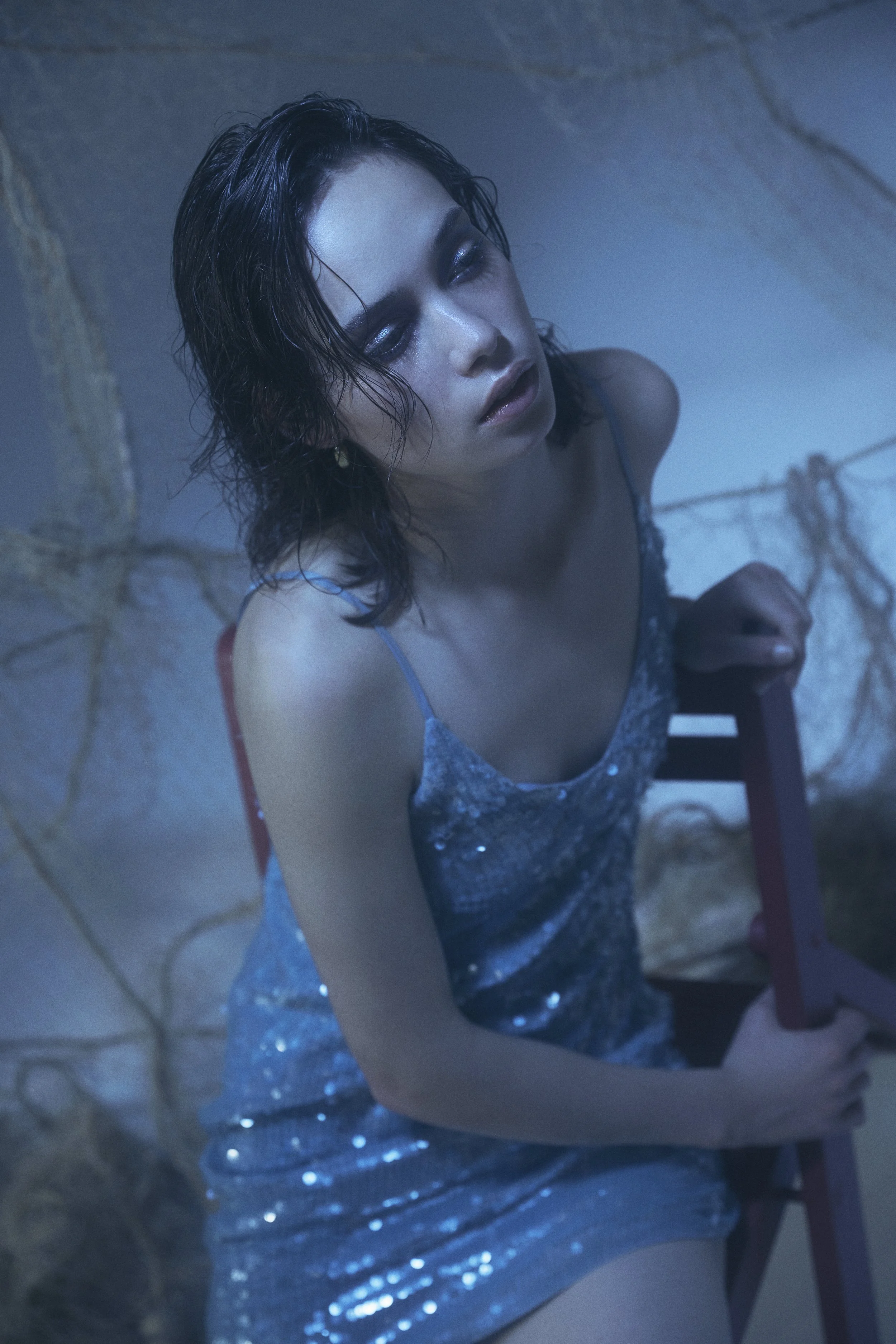 A woman with wet, dark hair and shiny makeup, wearing a sparkly blue dress, sitting on a small red chair, holding onto the backrest, with a background dimly lit with a blue hue.