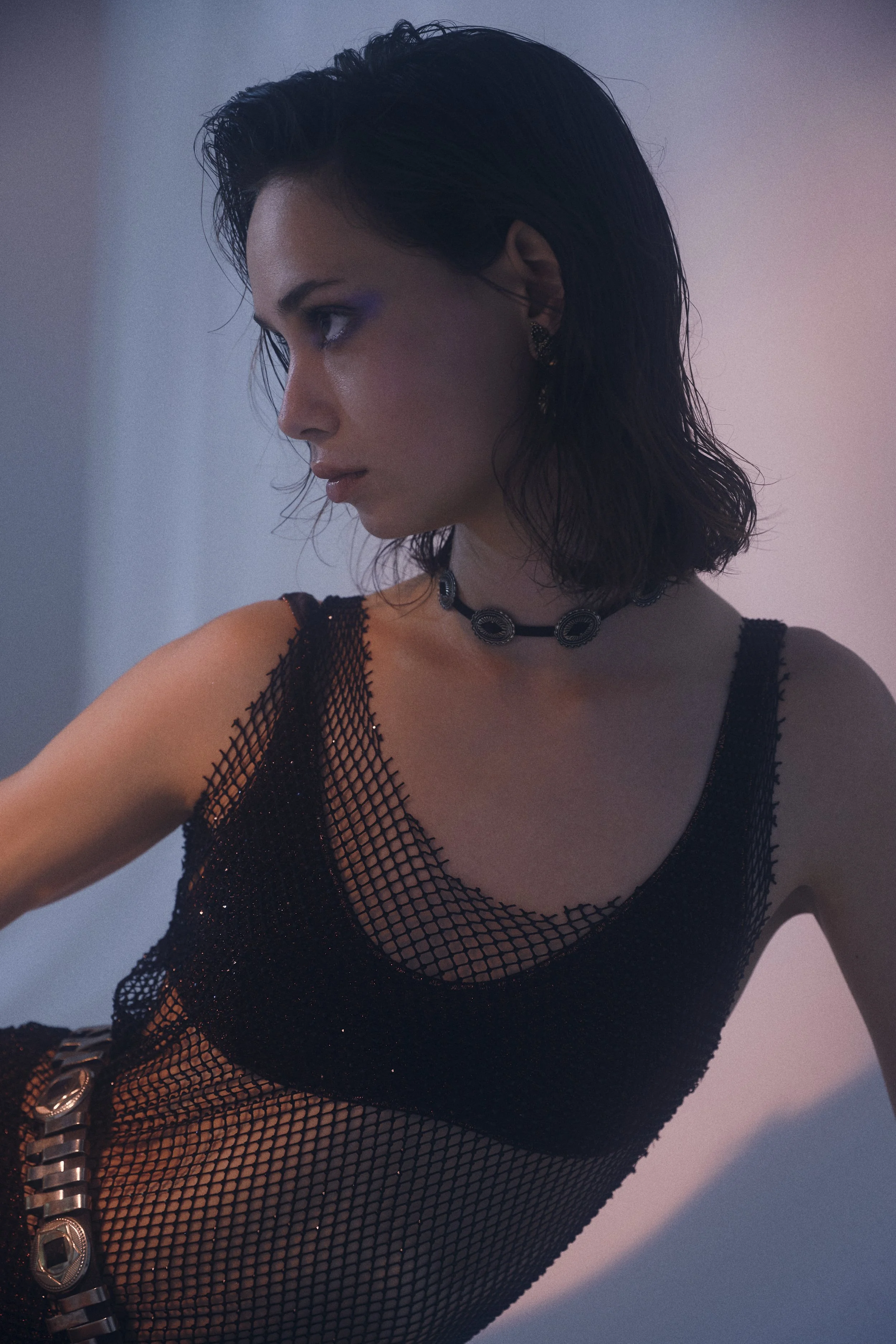 A woman with dark, wet hair is wearing a black fishnet top, a black choker necklace, and earrings, posing indoors with pink and blue lighting.