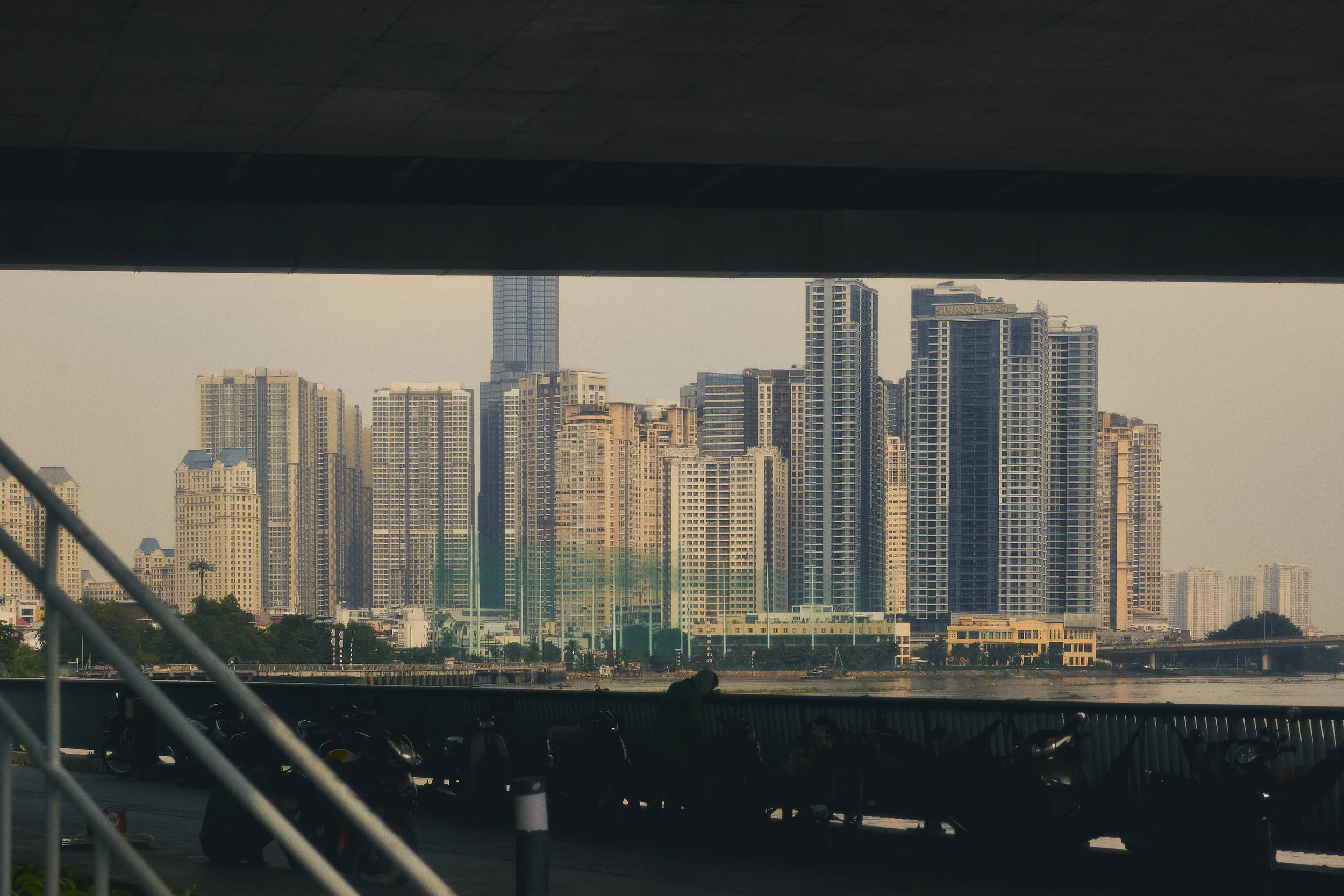 View of a city skyline with tall modern buildings seen from underneath a bridge, with a parking lot and motorcycles in the foreground.