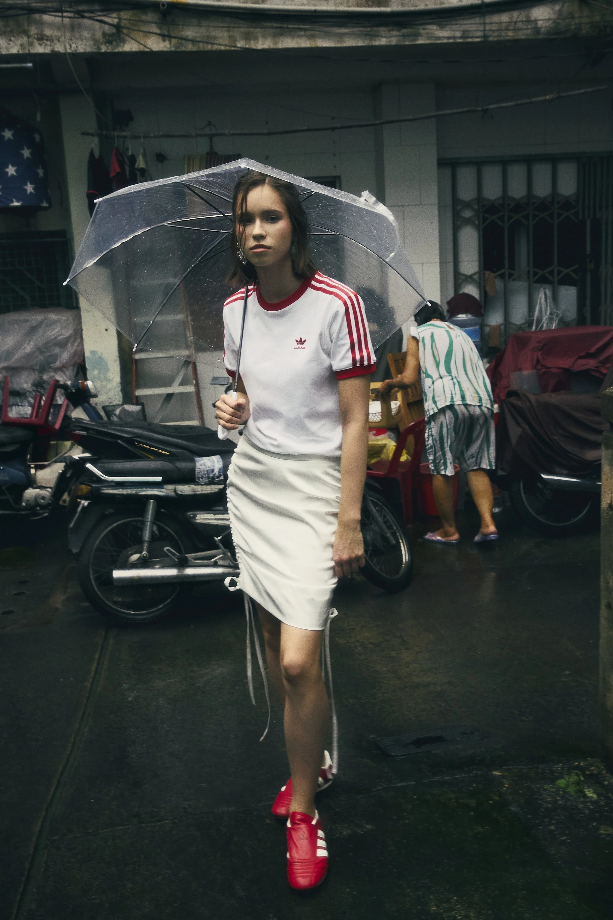 A woman wearing a white Adidas t-shirt, white skirt, and red shoes, holding a transparent umbrella in a street scene with motorcycles and people in the background.