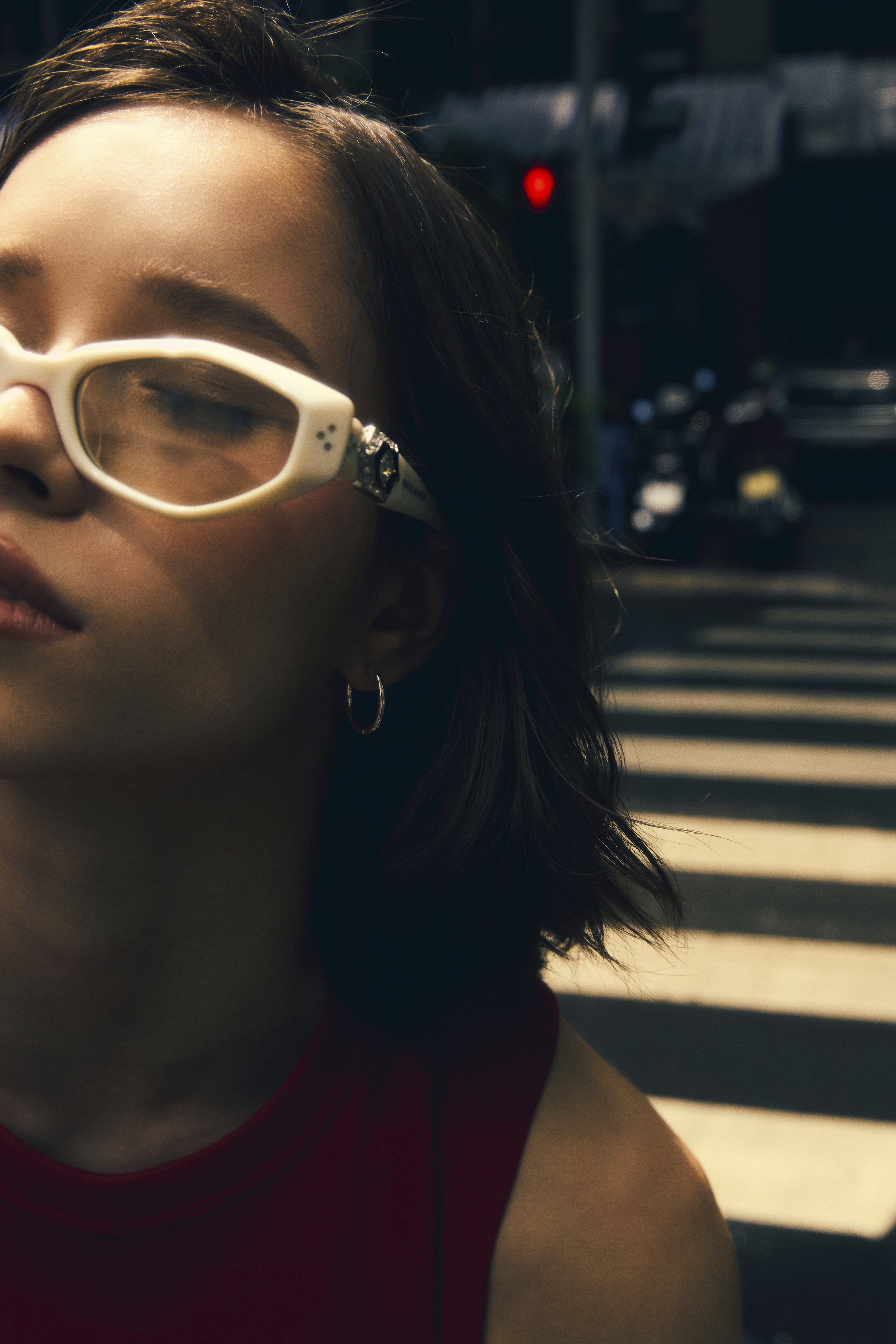 A woman with dark hair wearing white sunglasses and hoop earrings stands at a crosswalk on a city street during daytime.