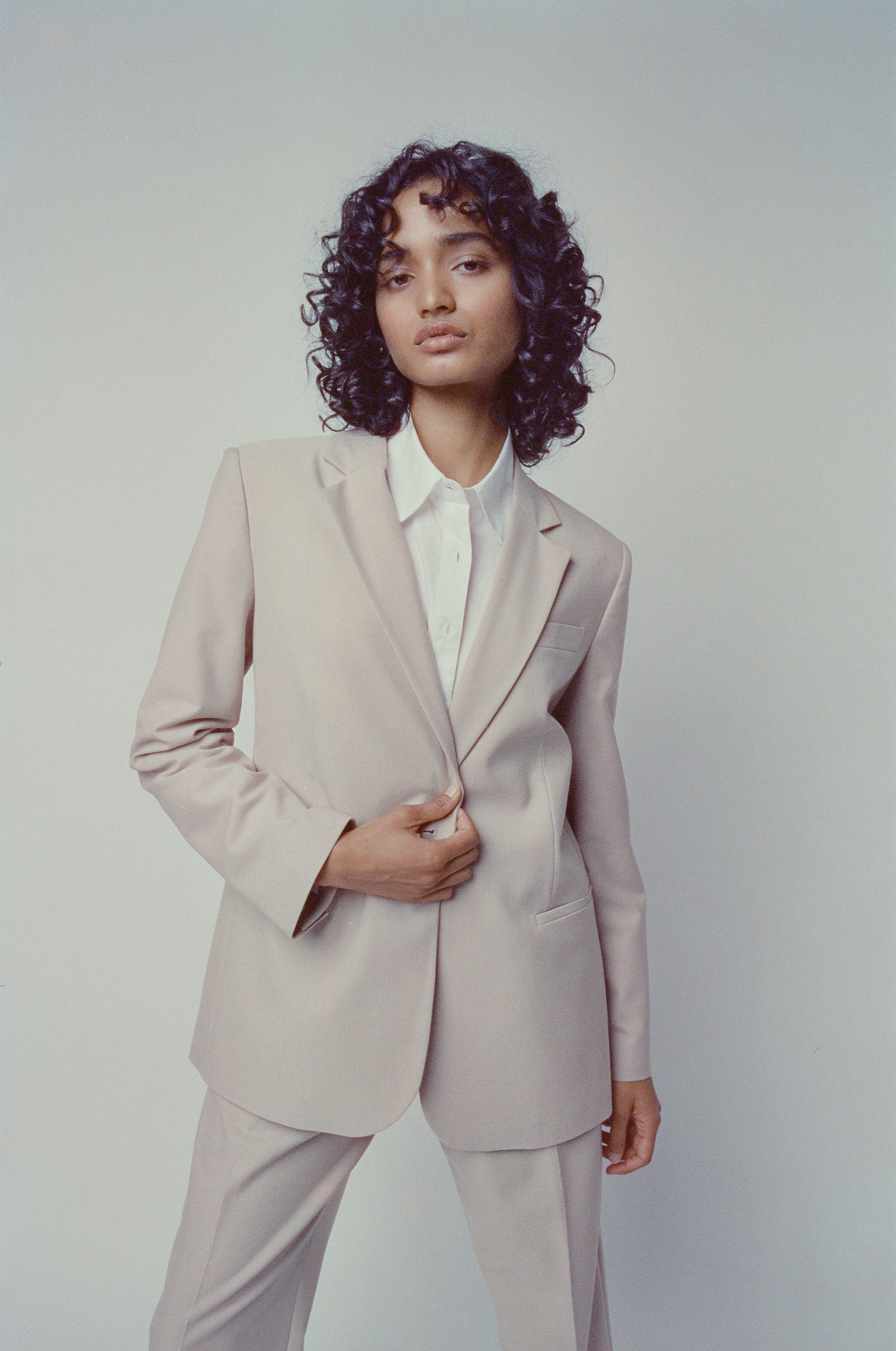 A woman with curly black hair wearing a beige suit and white shirt standing against a plain neutral background.