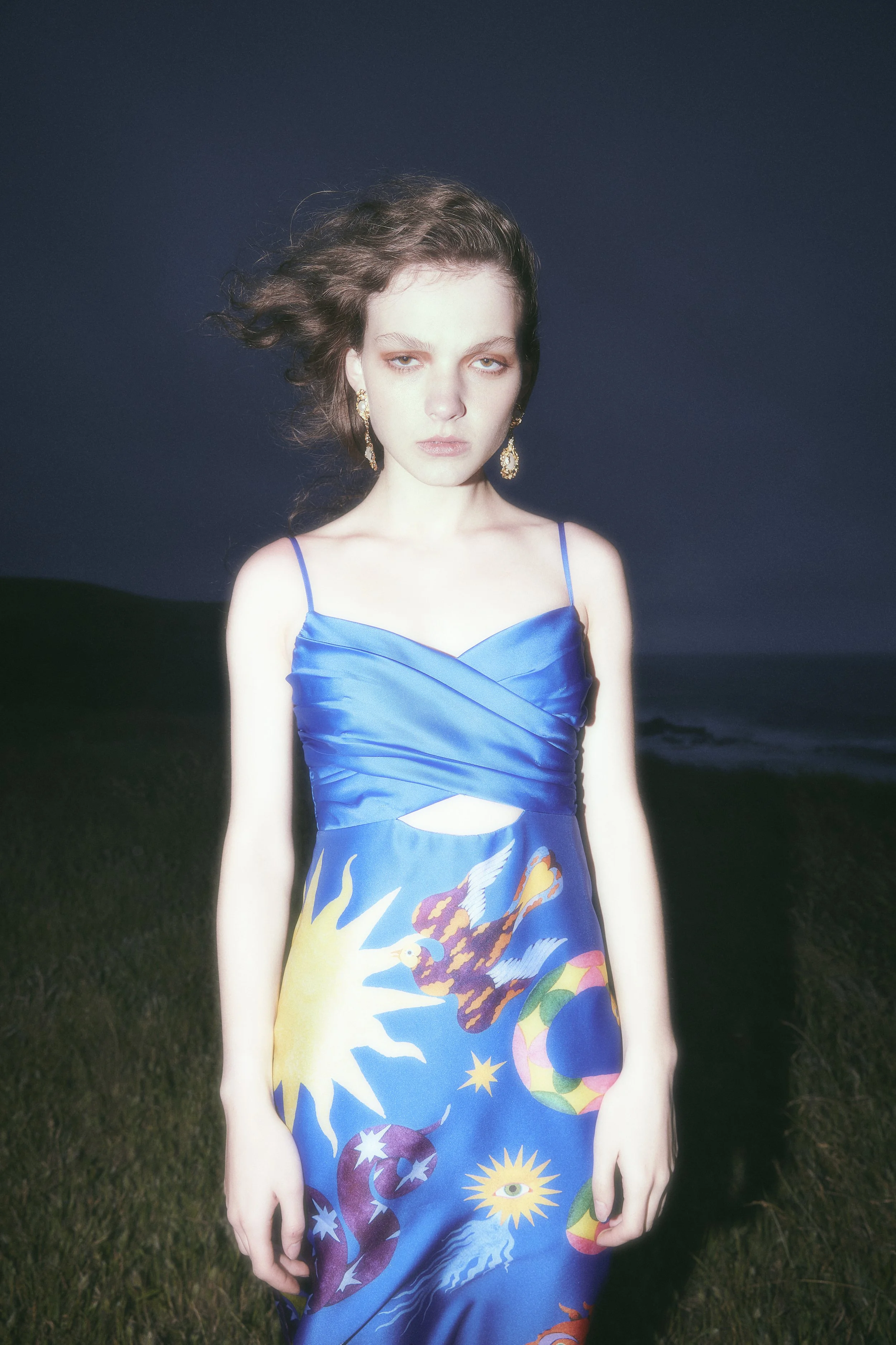 A woman with fair skin and dark wavy hair stands outdoors at night, wearing a blue dress with cosmic and celestial designs, and holding a serious expression.