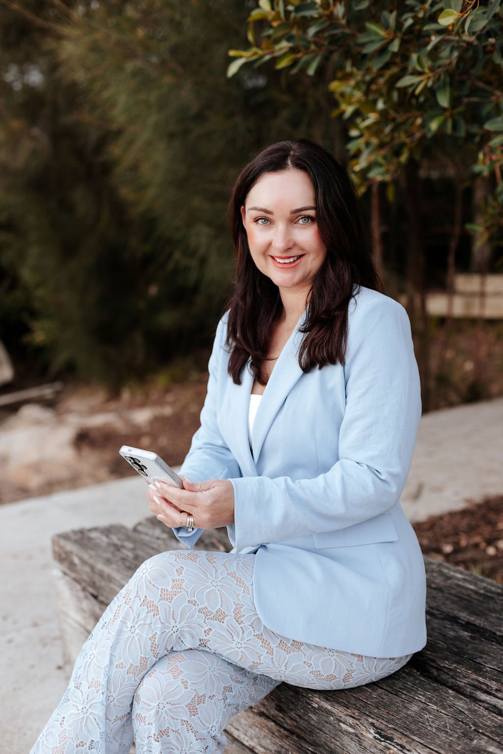 A woman with dark brown hair and blue eyes sitting on a wooden bench outdoors, smiling while holding a smartphone, wearing a light blue blazer and lace-patterned pants, with greenery in the background.