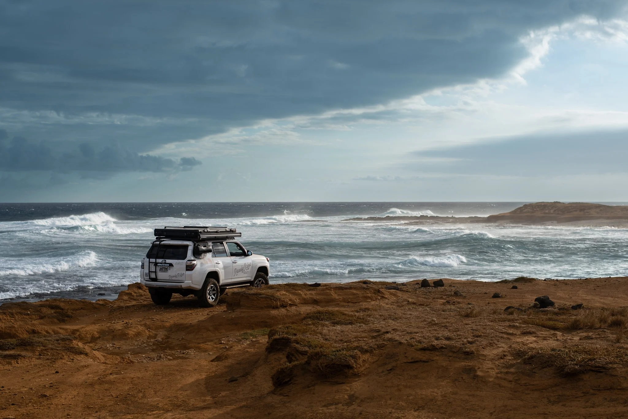 A white campervan parked on a rugged dirt cliff in hawaii overlooking the ocean with waves crashing and cloudy sky.