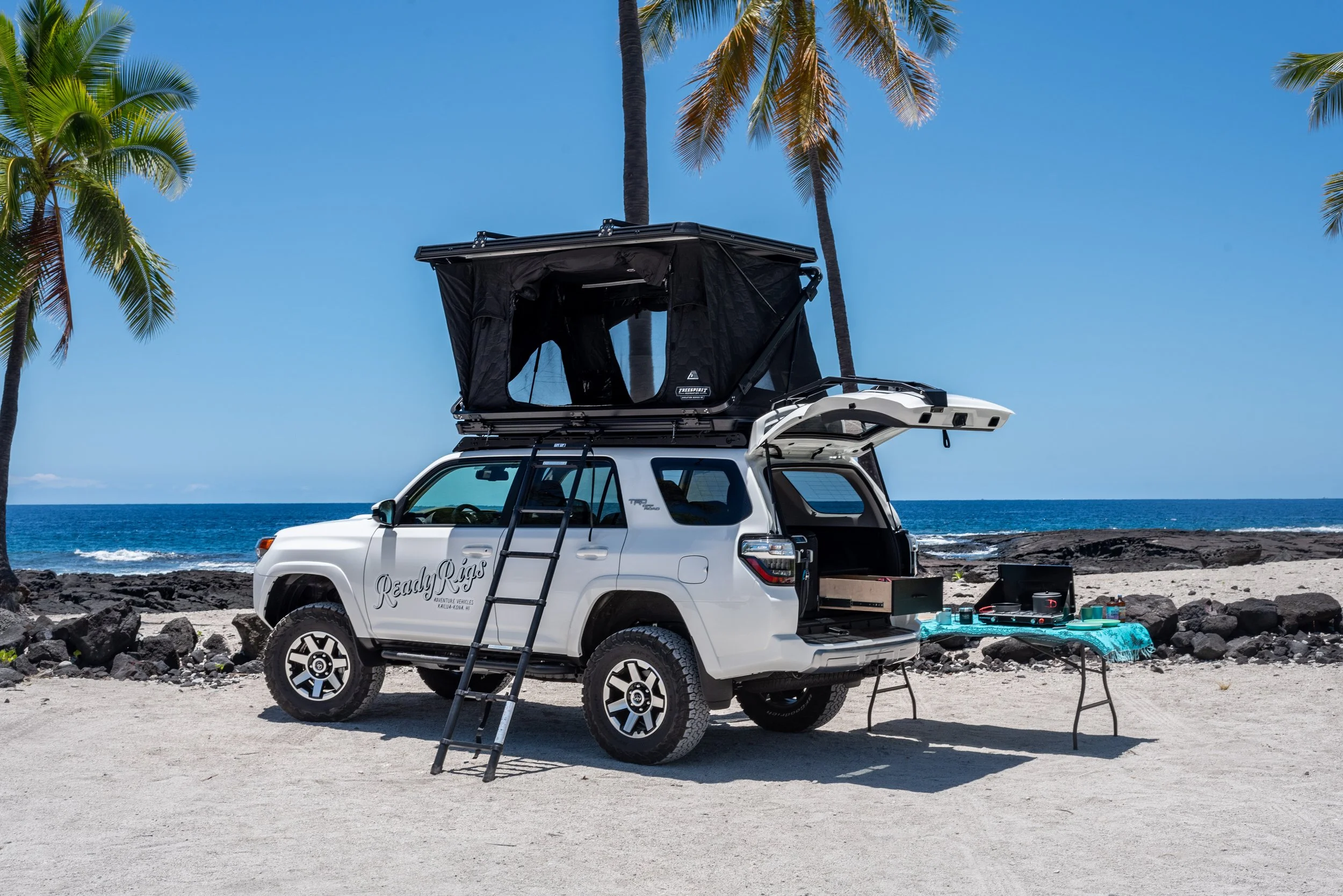 A white SUV with a rooftop tent and a ladder on a sandy beach, with palm trees, rocks, and ocean in the background, and outdoor gear on a table nearby.