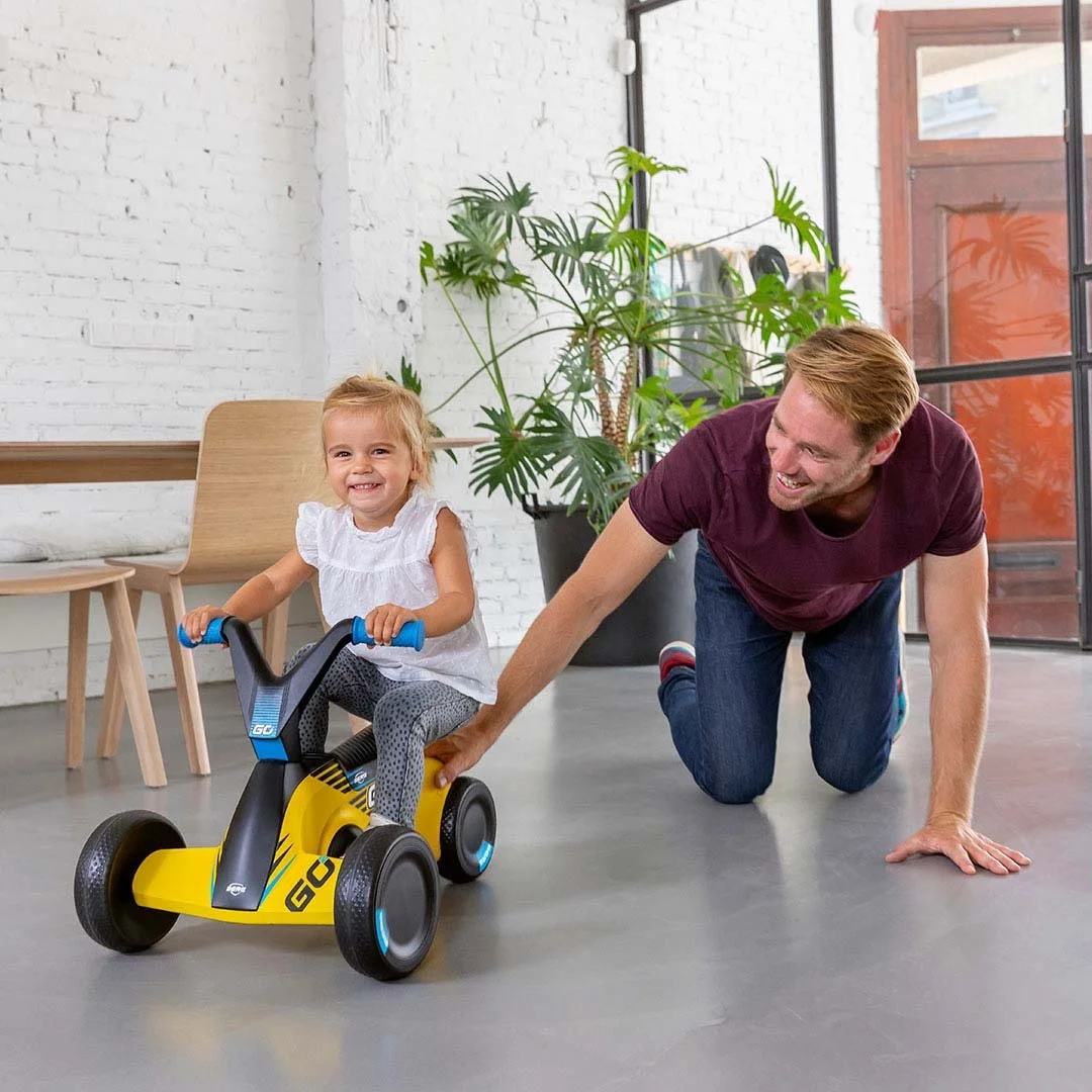 Ein lachendes kleines Mädchen fährt auf einem gelben Dreirad, während ein Mann auf dem Boden hinter ihr ihn schiebt, in einem modernen Wohnzimmer mit weißen Wänden und grünen Pflanzen.
