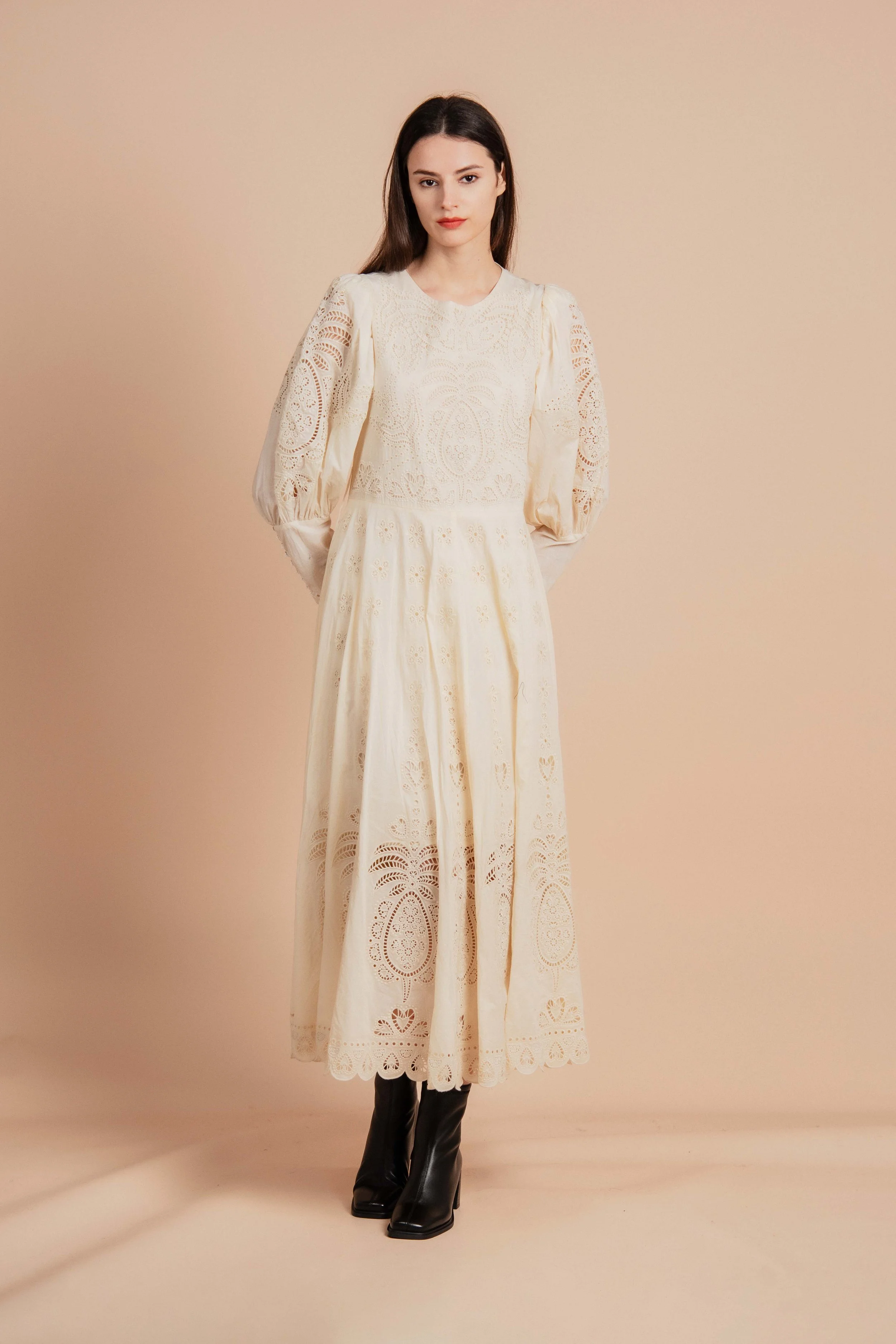 A woman wearing a long, cream-colored lace dress with puffed sleeves and black boots standing against a beige background.