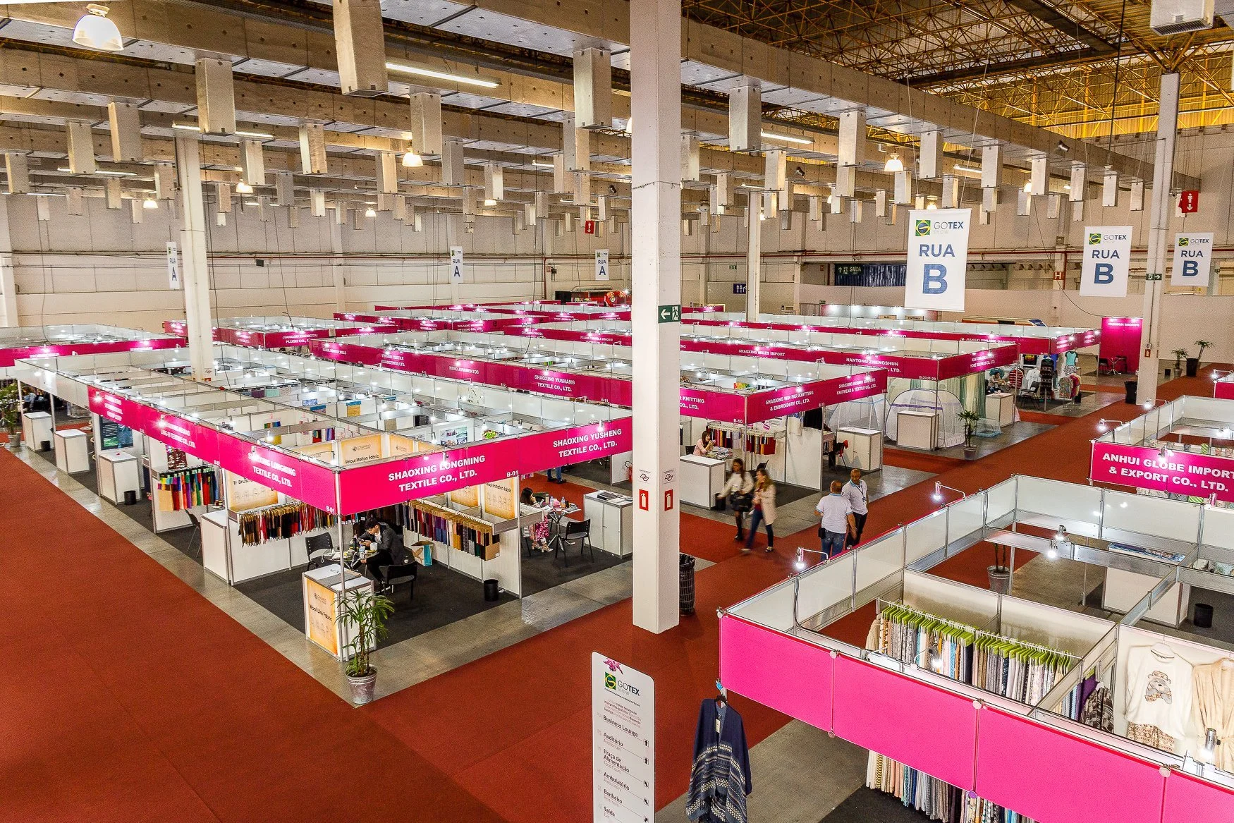 An indoor trade show with multiple booths featuring pink signs, some with textiles and fabrics. The ceiling is high with visible metal beams and numerous hanging lights. People are walking around, visiting booths, and exploring the exhibits. Large signs indicate the area as 'RUA B' and there is a sign for the company GOTEX. The overall setting appears spacious and organized.